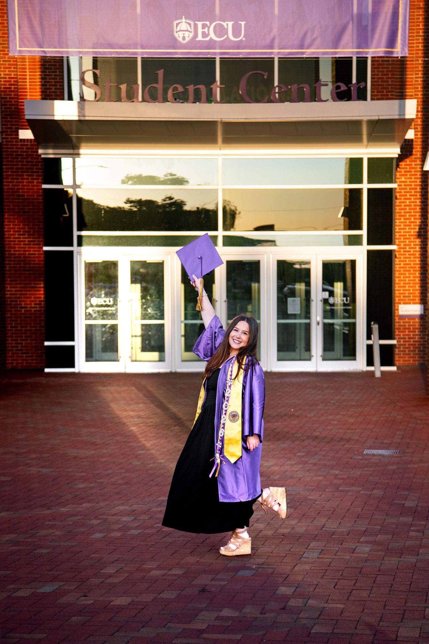 Excited graduate in purple ECU cap and gown tosses mortarboard in front of Student Center.