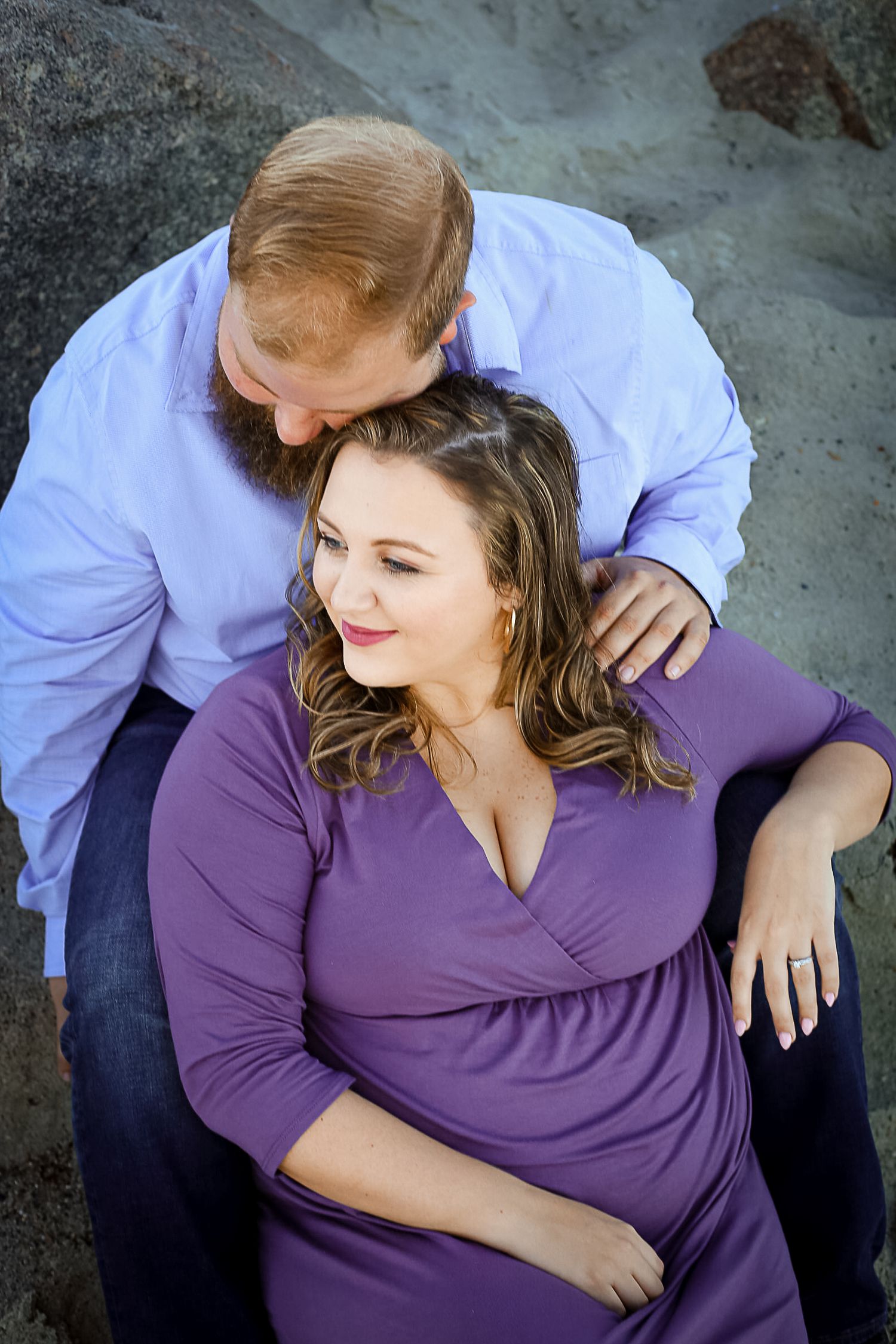 A couple shares a tender moment, man in blue shirt kissing woman's head as she smiles in a purple dress outdoors.