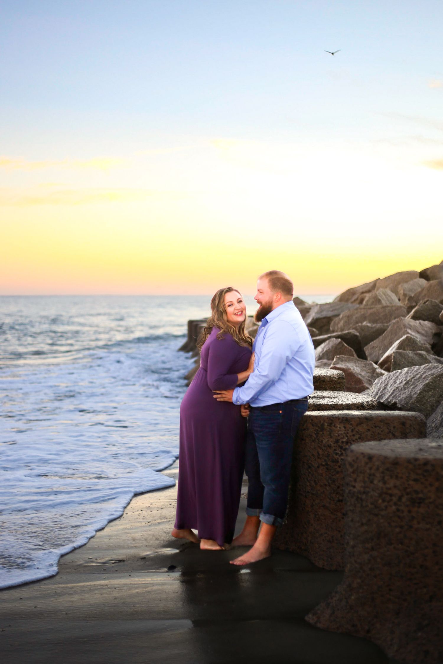 Couple sharing a romantic moment on a rocky beach at sunset, woman in purple dress with baby bump.