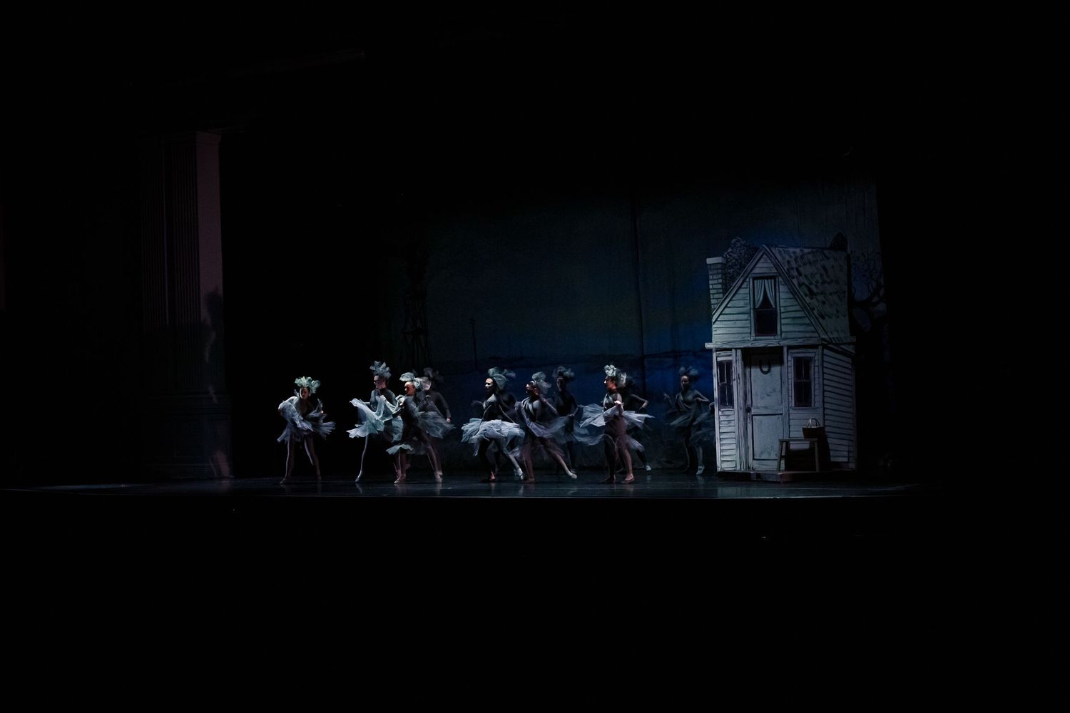Ballet dancers perform on a dimly lit stage beside a miniature house set during a theatrical production.