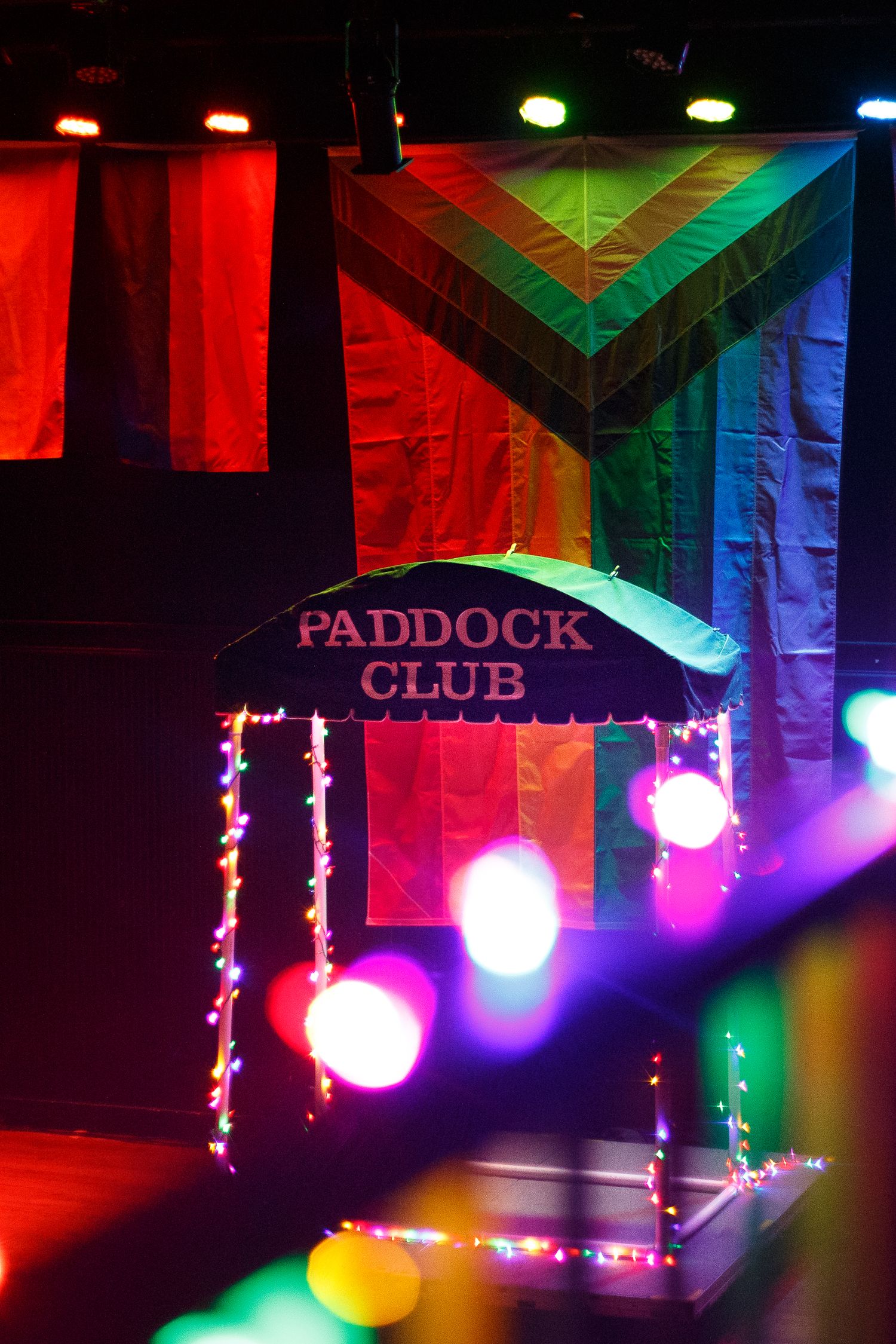 Paddock Club sign with rainbow pride flag backdrop and colorful string lights on a dark stage.