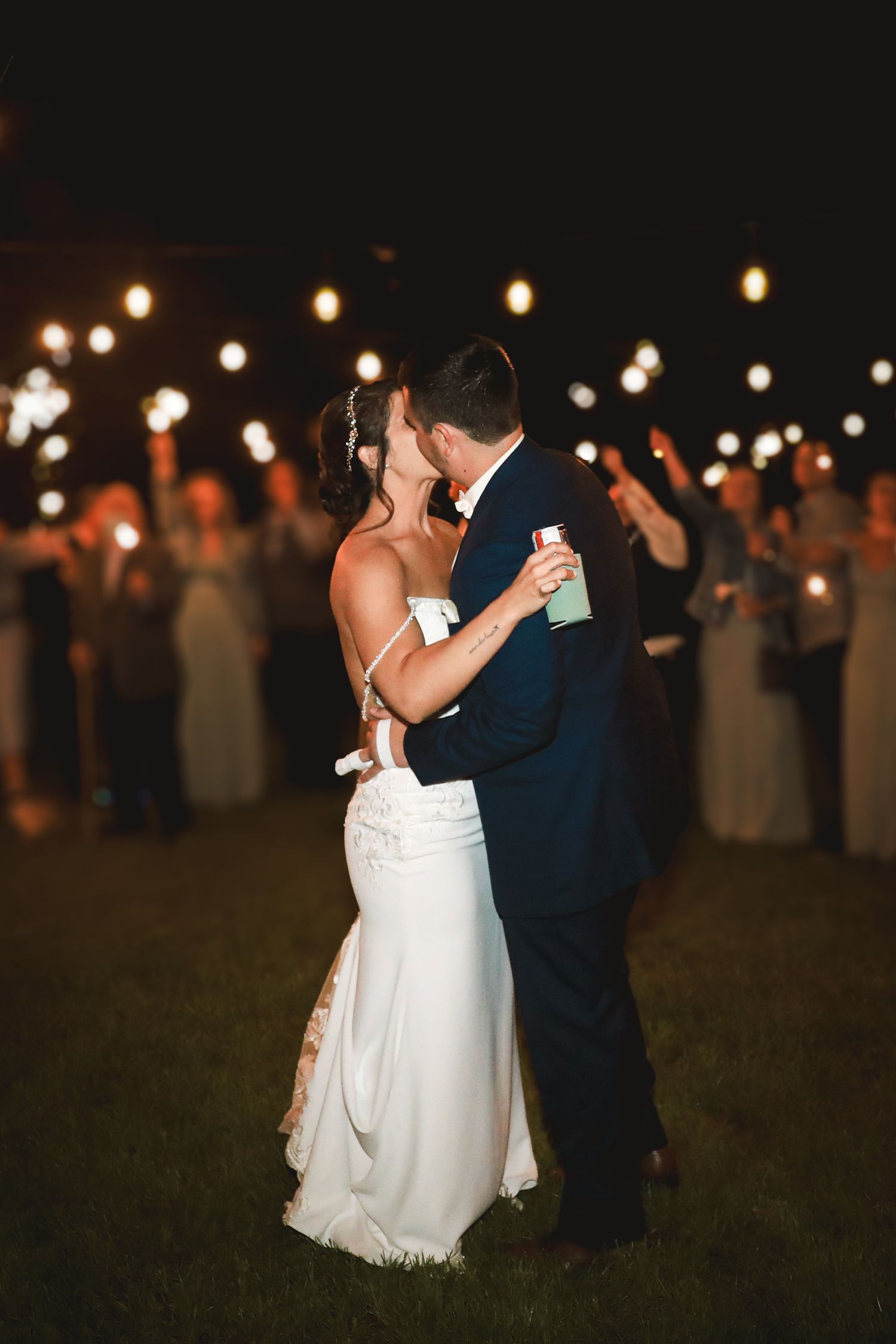 Bride and groom share a romantic kiss surrounded by guests holding sparklers at a nighttime wedding reception.