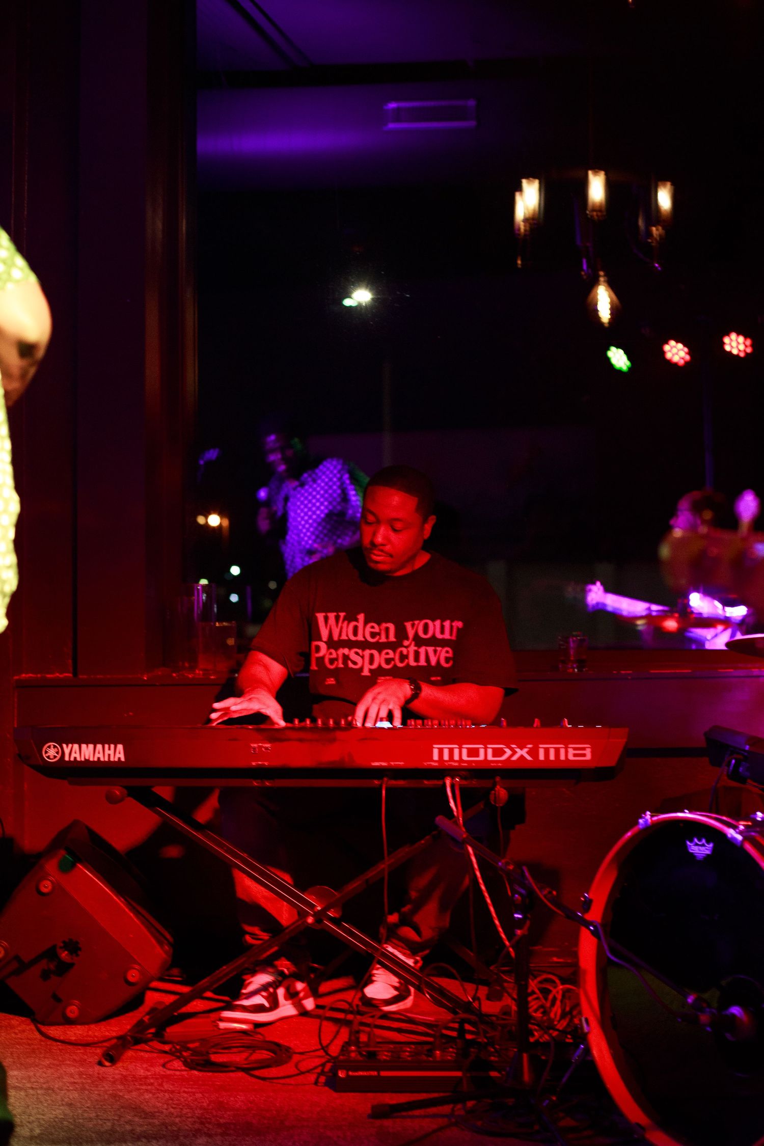 Musician playing a Yamaha MODX8 keyboard on a dimly lit red-lit stage wearing a “Widen Your Perspective” shirt.