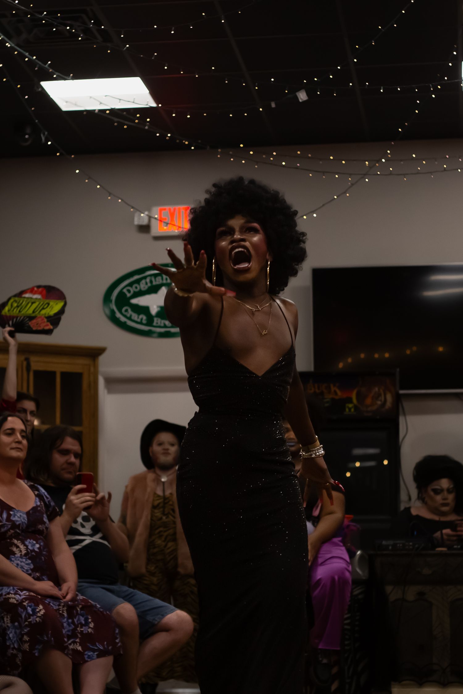 Performer in black sequin dress with afro hairstyle singing passionately at an indoor drag show event.