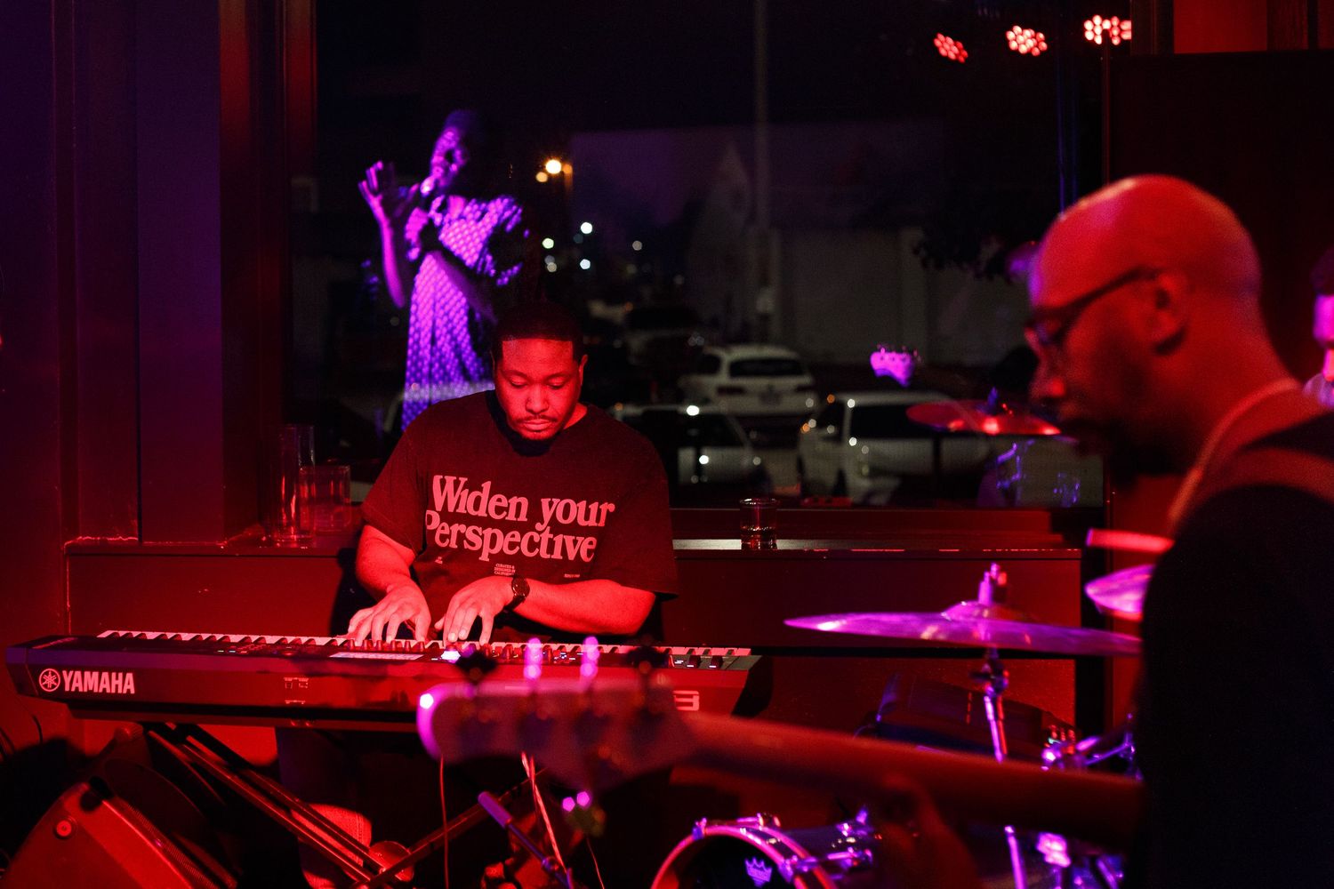 Live band performance with keyboard player and drummer on stage, bathed in red light, with singer in background.