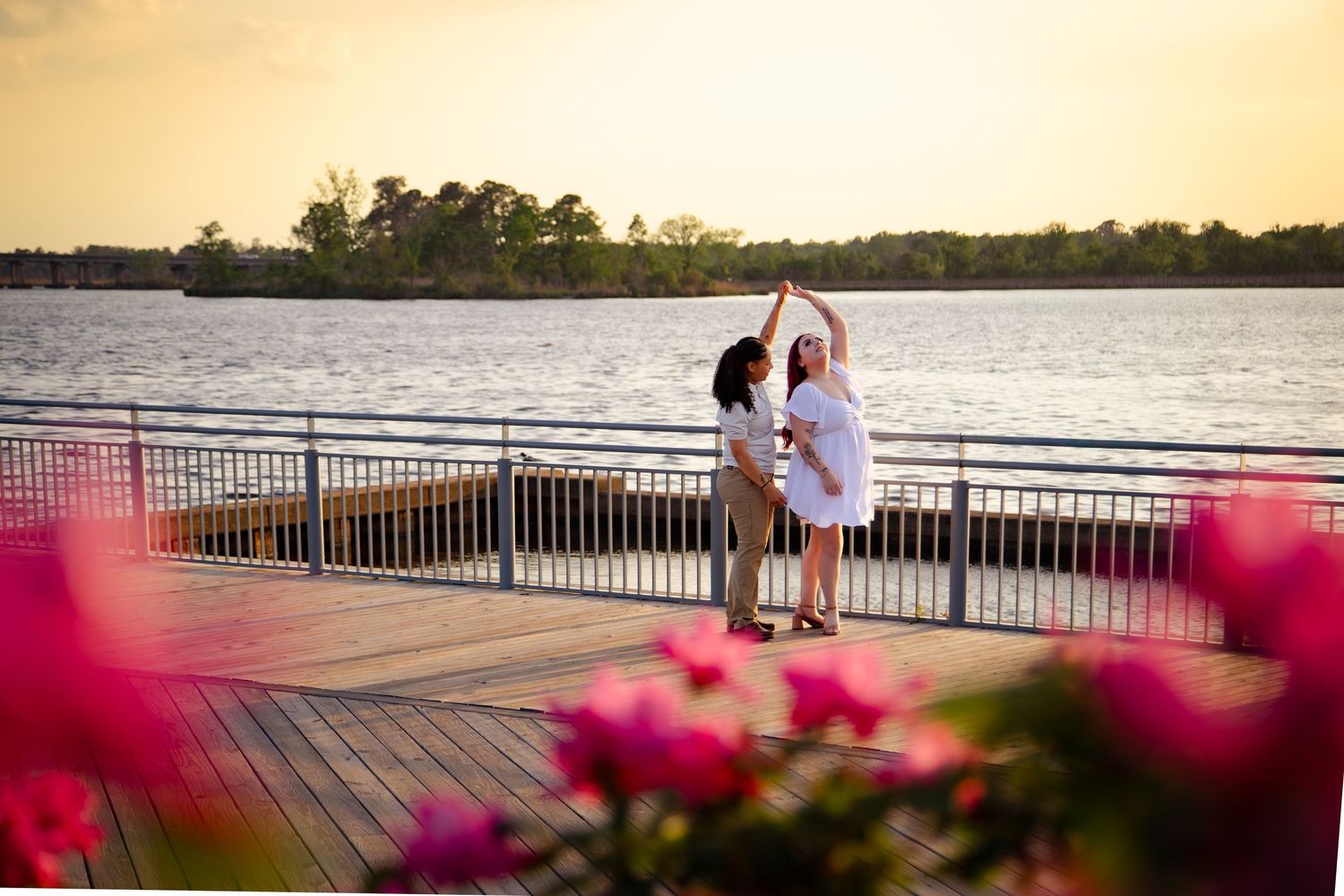 Couple dancing on a waterfront deck at sunset with pink flowers in the foreground and trees reflecting on calm water.