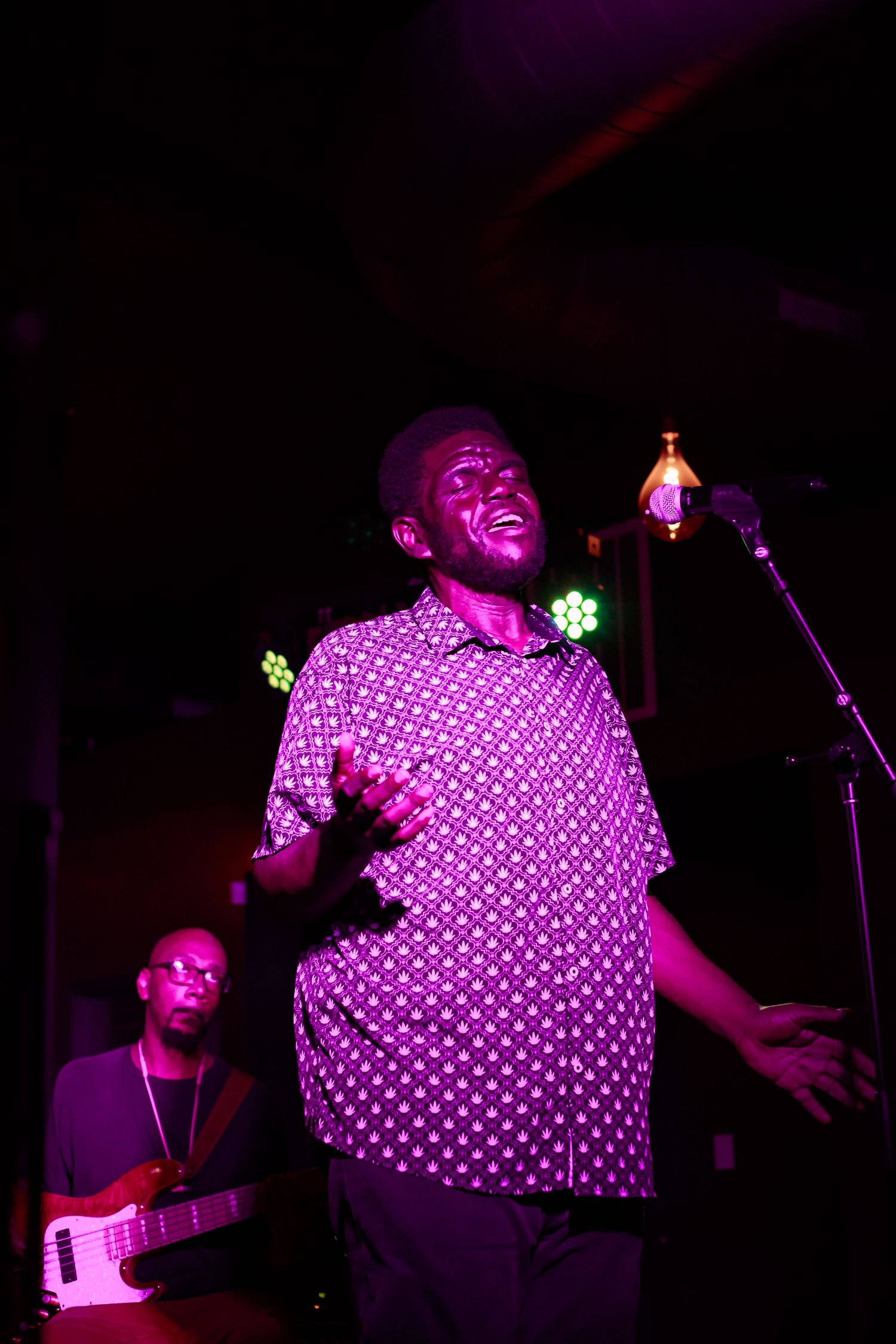 A singer performs passionately at a microphone under purple stage lights, with a guitarist visible in the background.