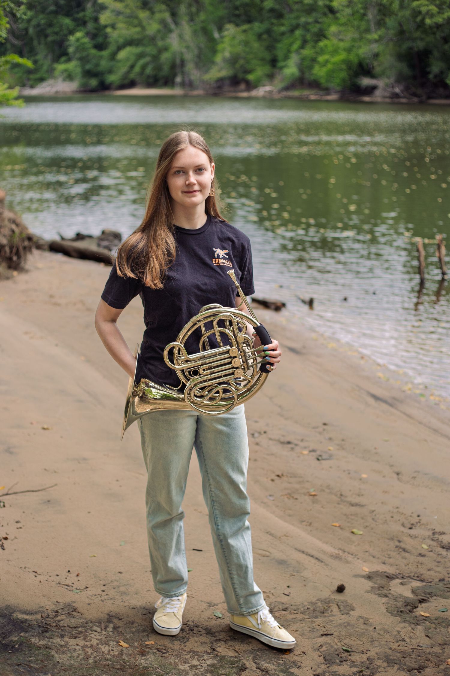 Young woman holding a French horn stands on a sandy riverbank with lush green trees in the background.