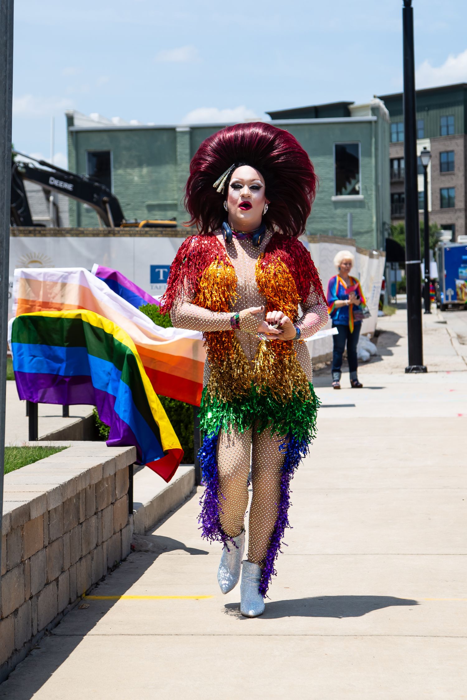 Drag performer in rainbow fringe costume and feathered headpiece walks proudly at an outdoor Pride parade event.