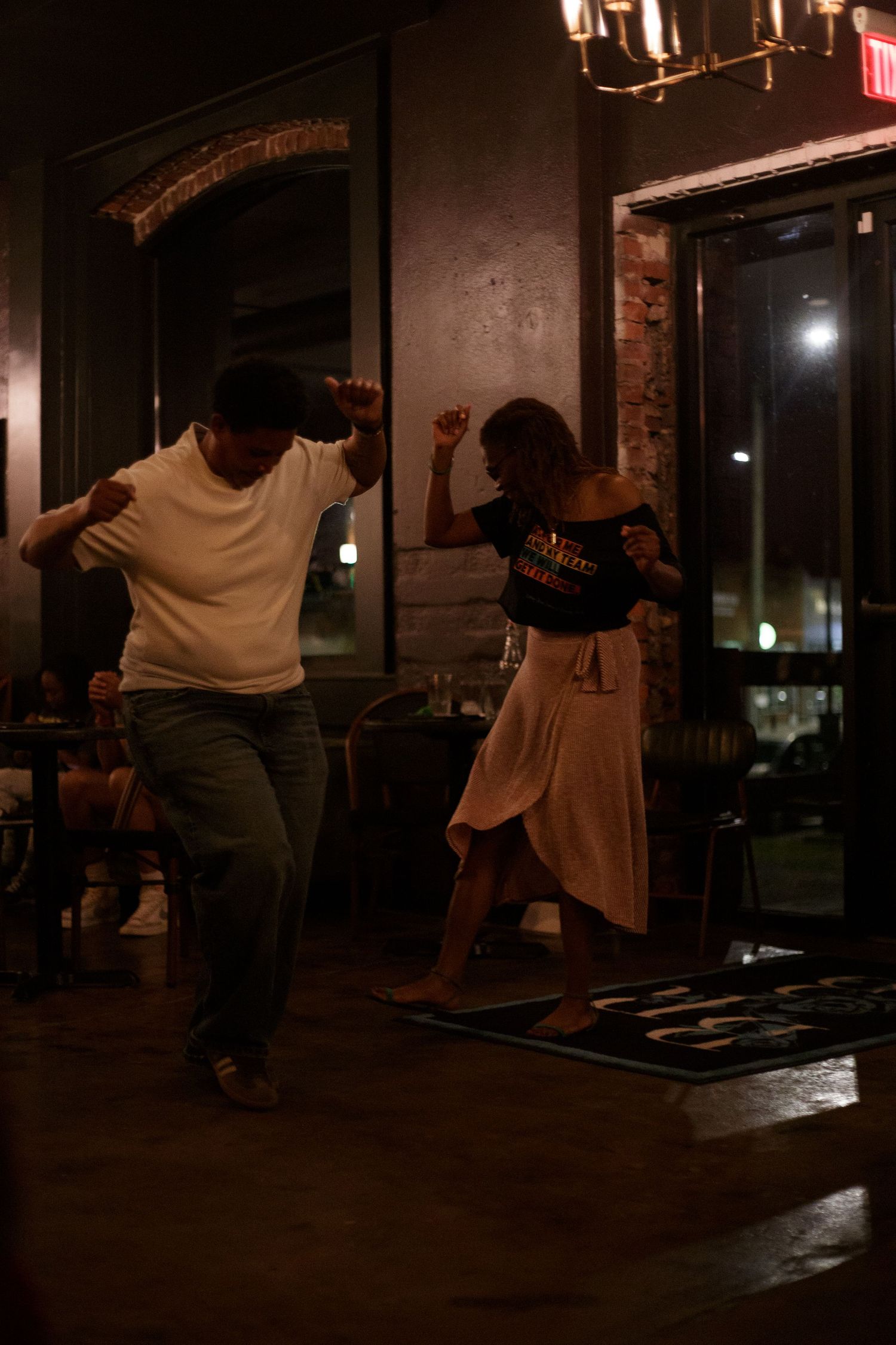 Two people dancing energetically inside a dimly lit bar or restaurant at night.