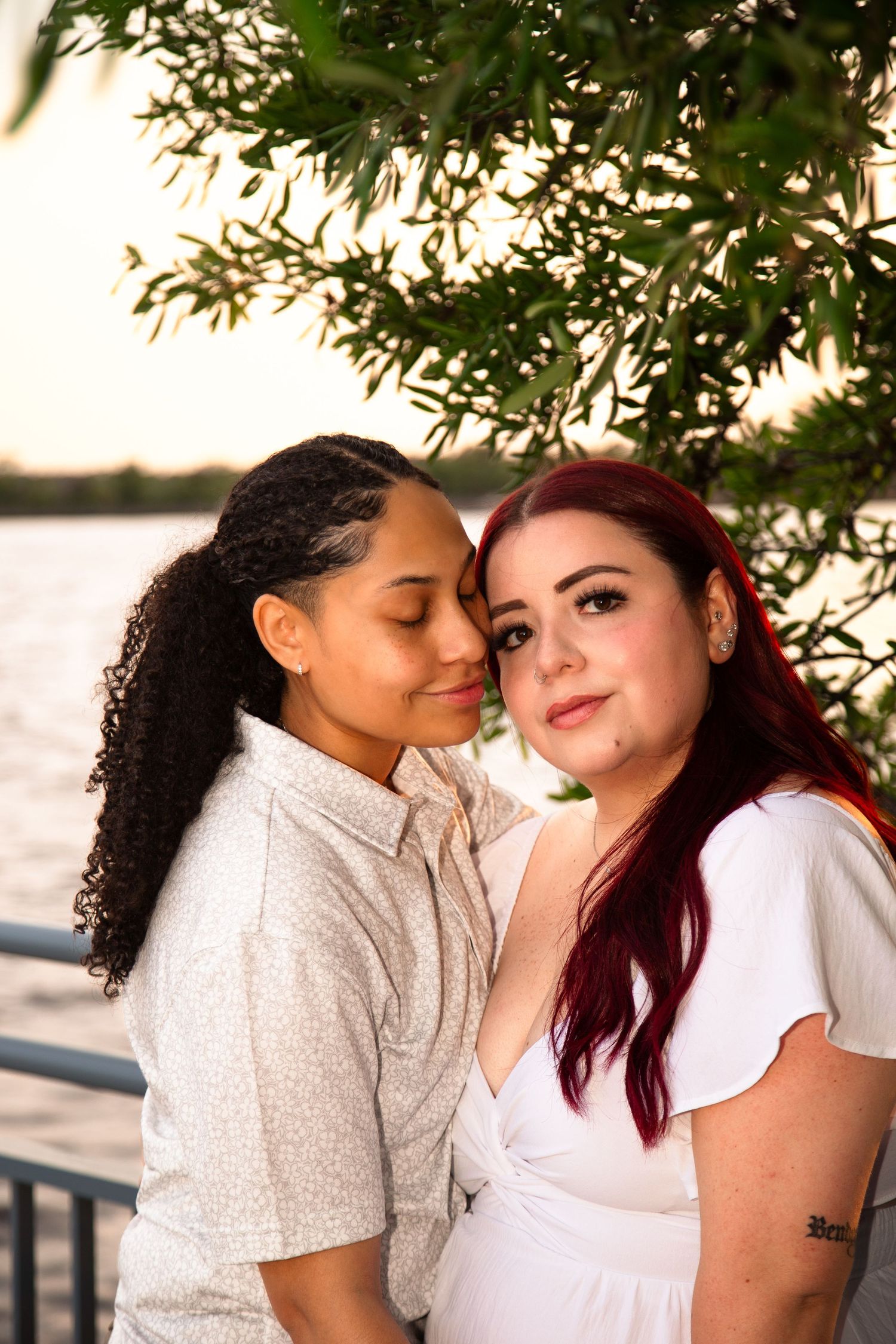 Two women embracing near waterfront at golden hour under willow tree.