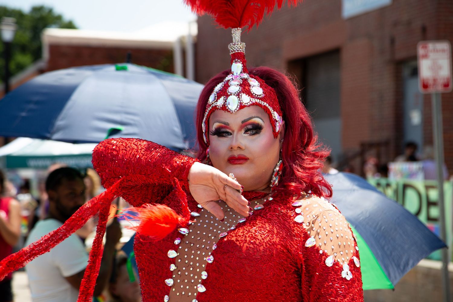 Drag queen in red sequin costume and feathered headpiece poses at outdoor Pride parade event.