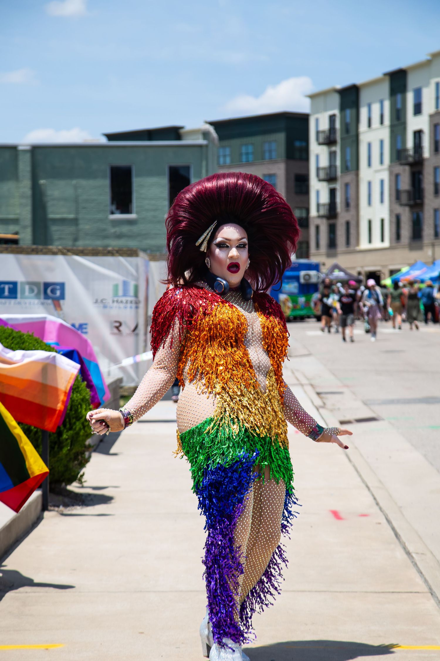 Drag performer in rainbow fringe dress and large red hair at Pride festival outdoors.
