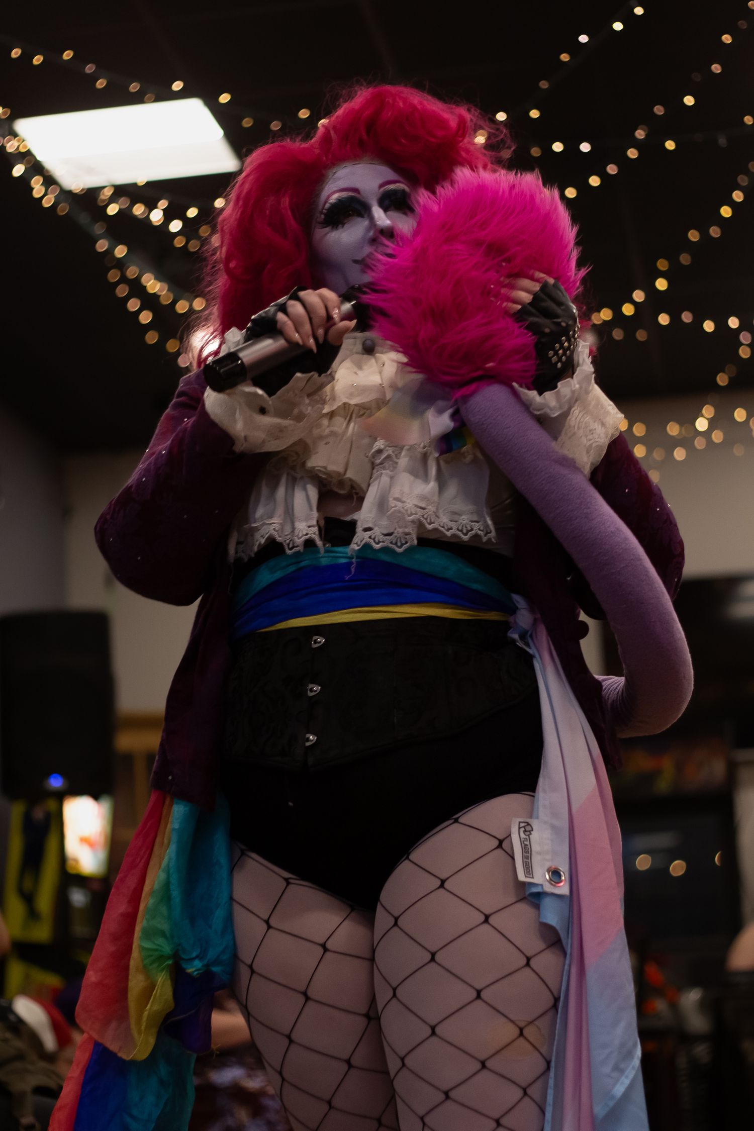 Drag performer in pink wig, corset, fishnet tights, and colorful costume holds microphone on stage.