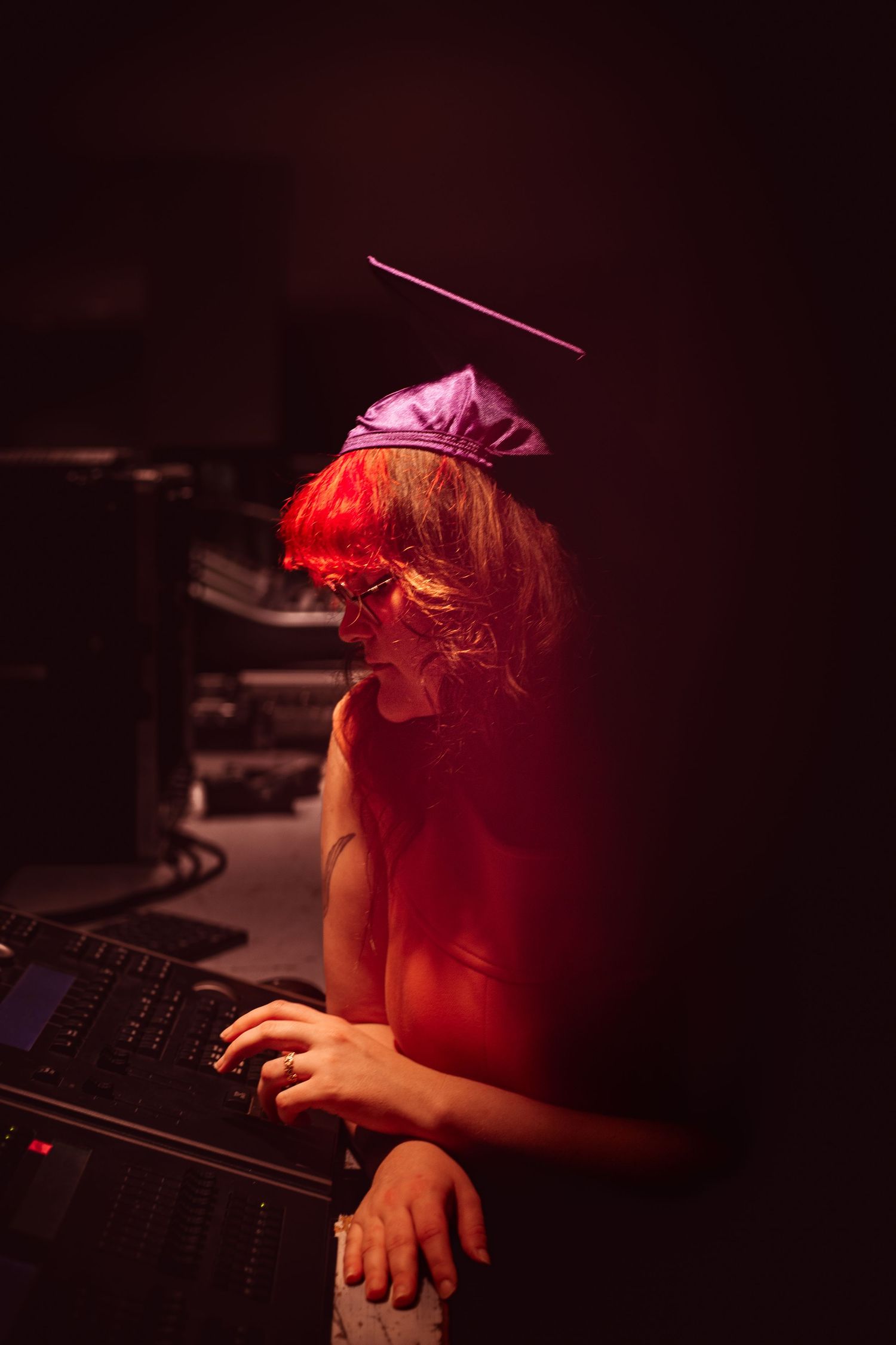 A DJ wearing a graduation cap operates a mixing console under dramatic red lighting in a dark venue.