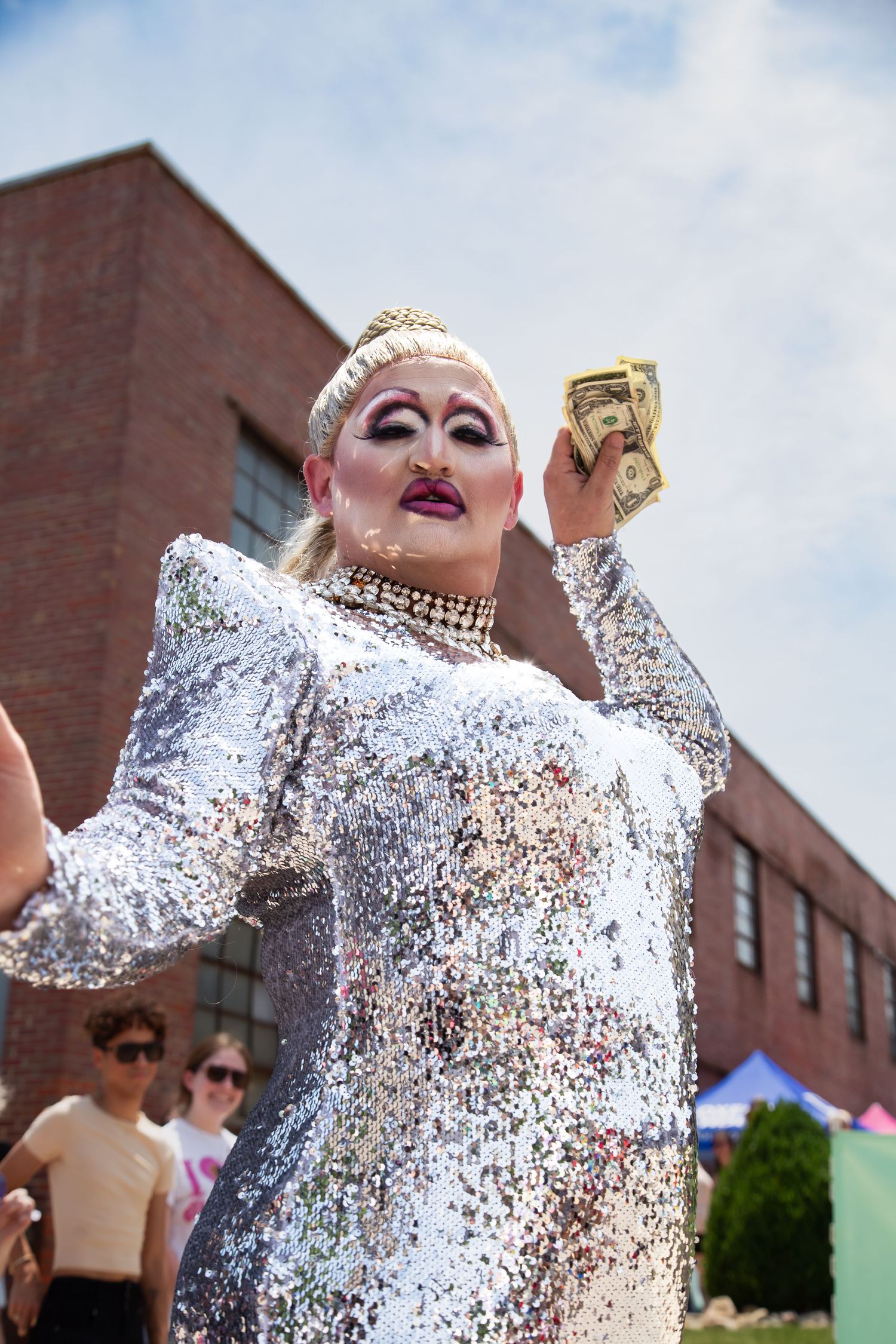 Drag queen in silver sequin dress holds dollar bills at outdoor Pride event.