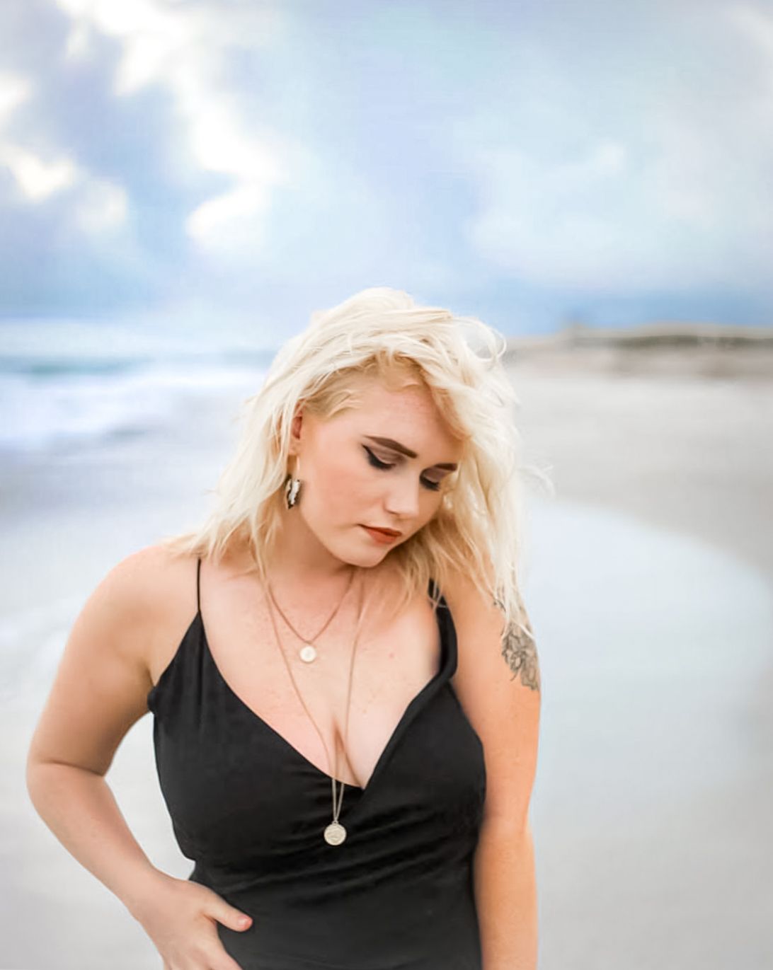 Blonde woman in black dress with tattoo poses on beach with ocean and cloudy sky backdrop.