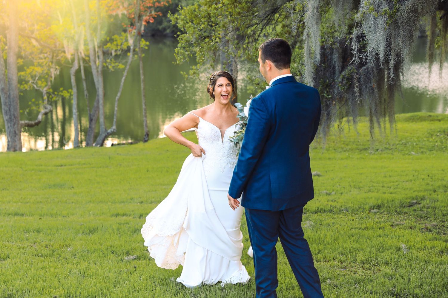 Bride in white gown and groom in blue suit share a romantic moment on lush green lawn near a scenic pond.