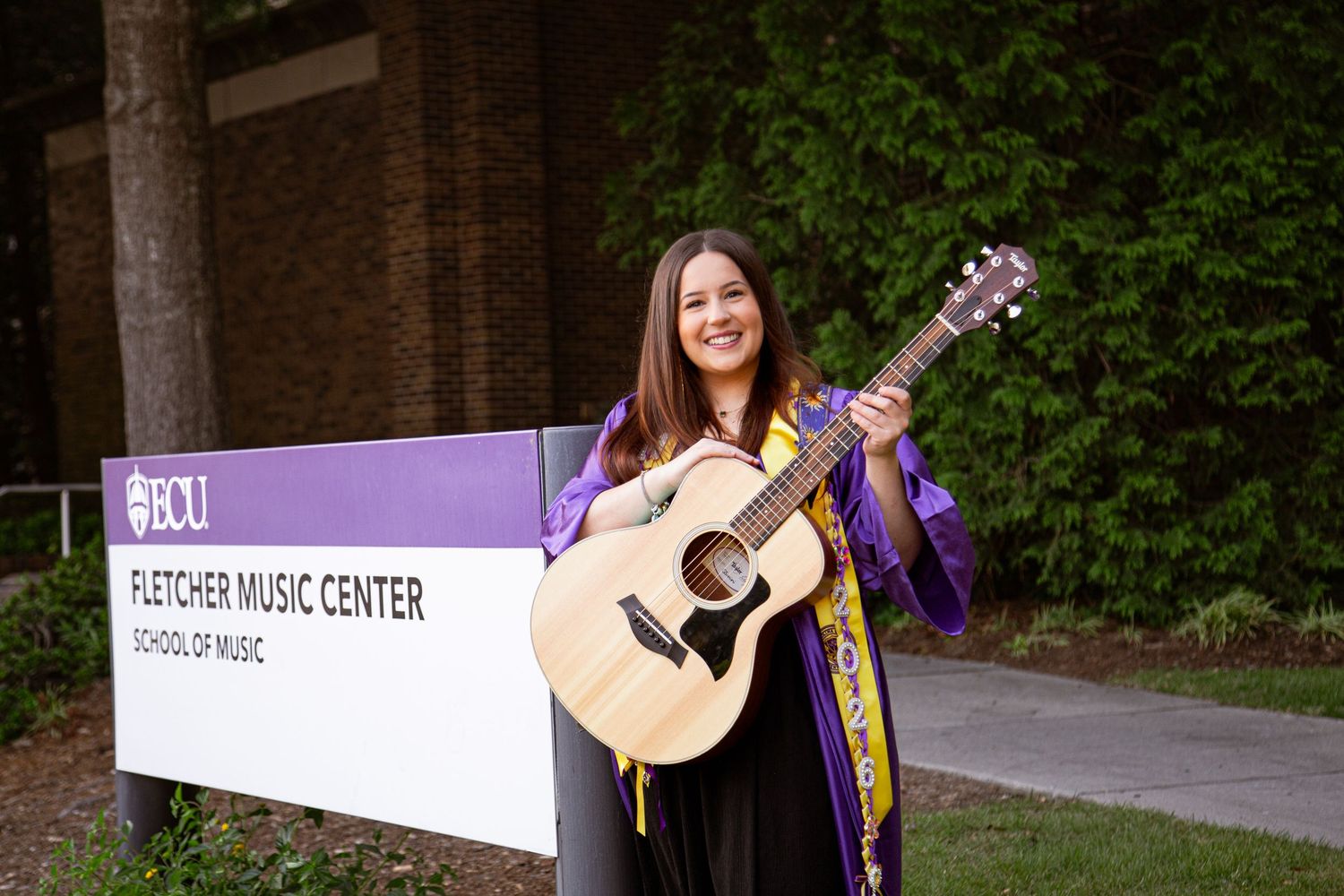 Smiling graduate holding acoustic guitar outside Fletcher Music Center ECU.
