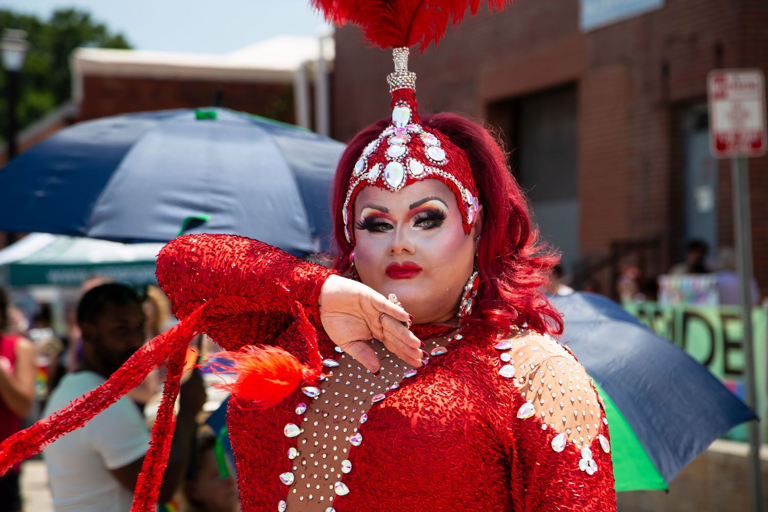 Drag queen in red sequin gown and feathered headdress performing at an outdoor Pride festival.