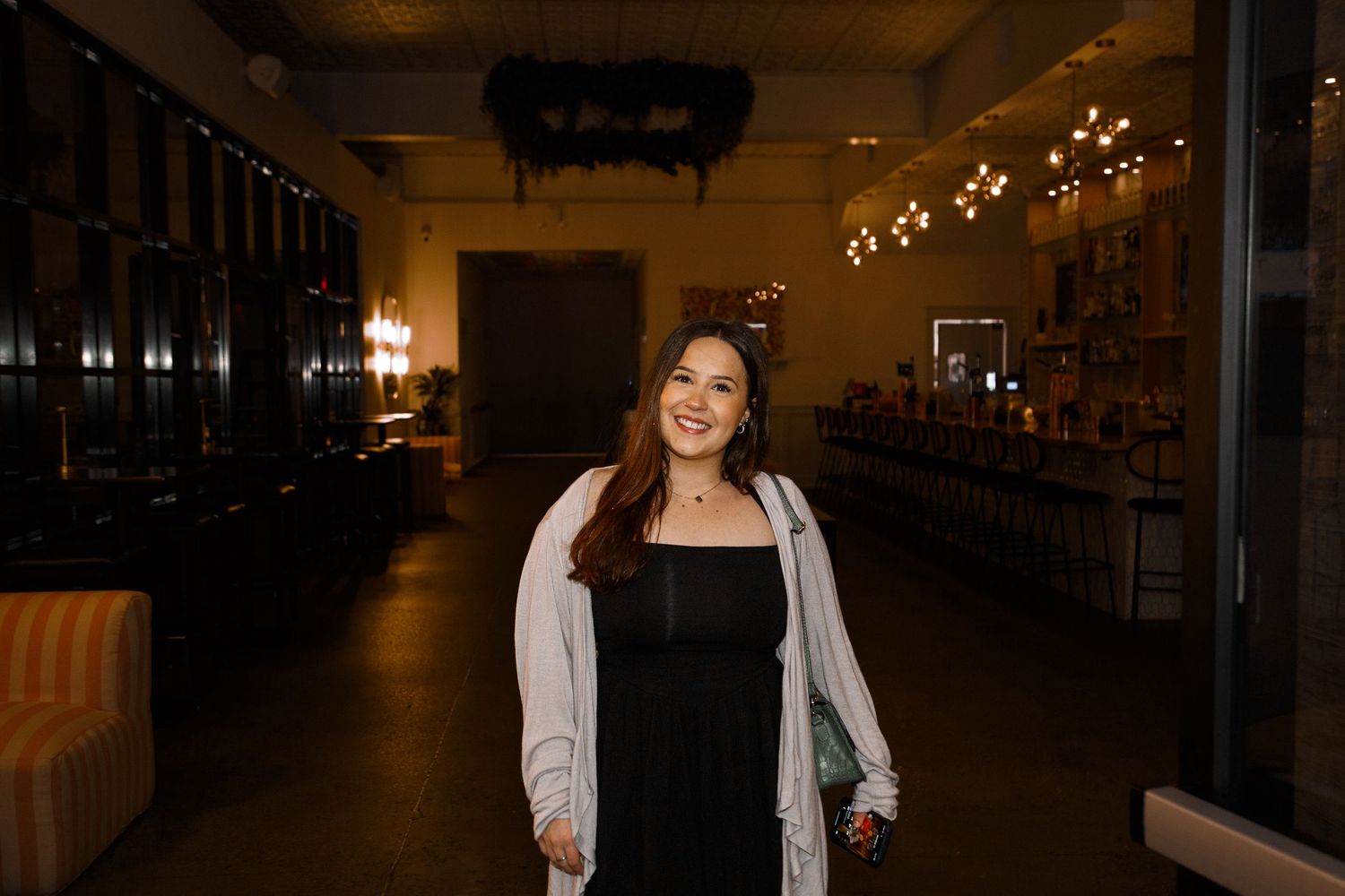 Smiling young woman in black dress and white cardigan standing in a warmly lit elegant venue interior.