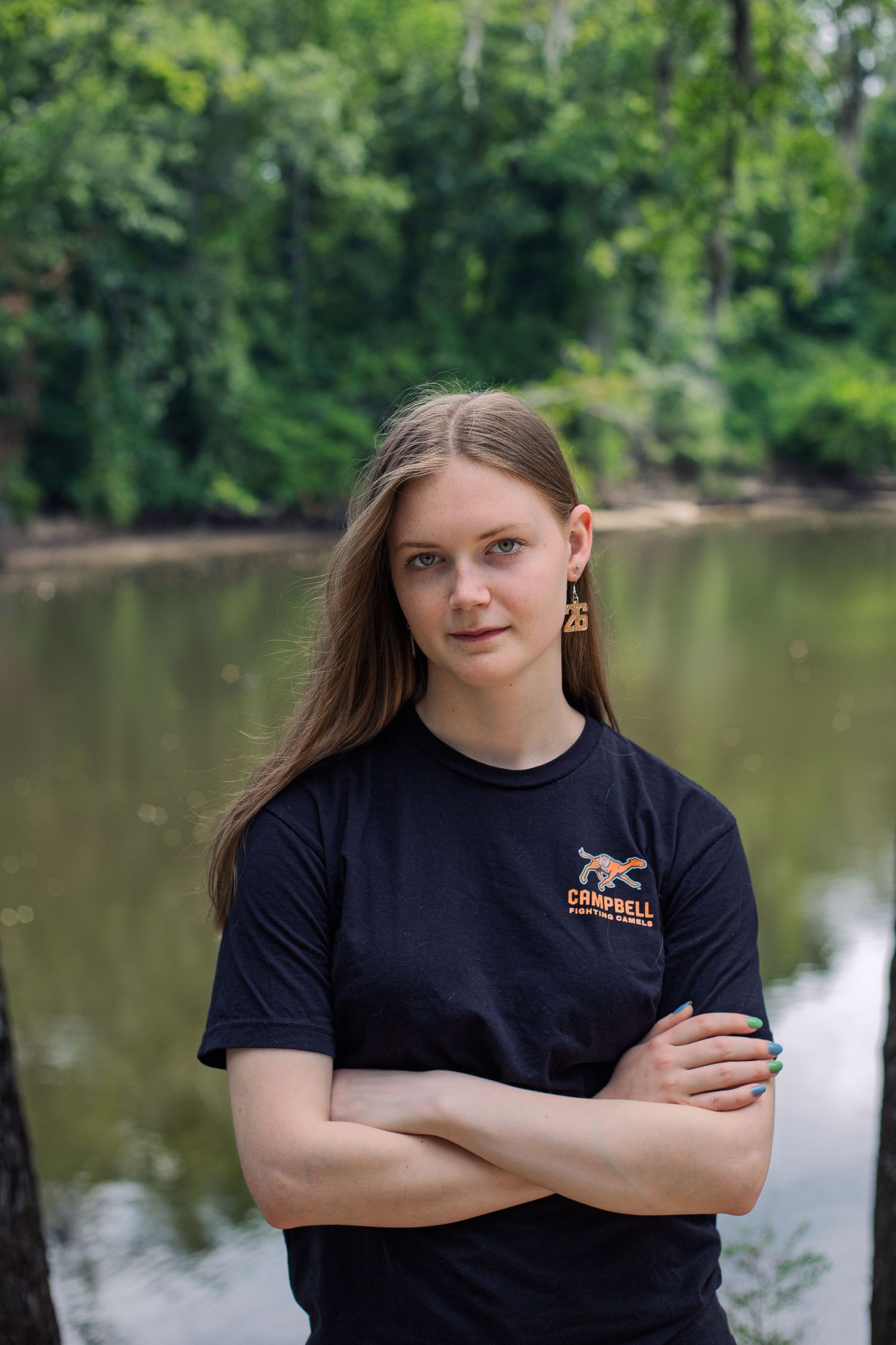 Young woman in Campbell t-shirt stands arms crossed by a serene river surrounded by lush green trees.