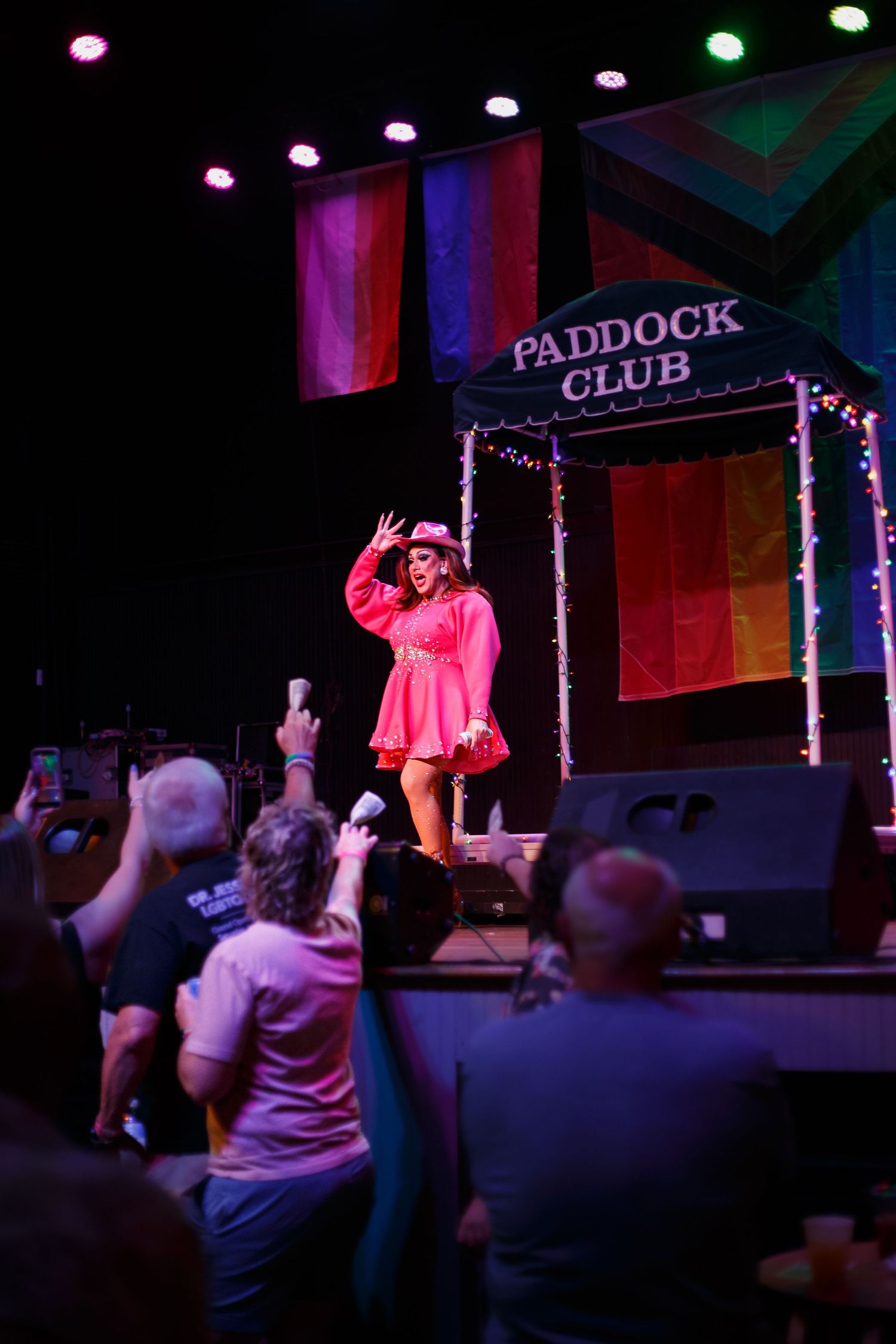 Drag performer in pink dress waves to crowd at Paddock Club stage with rainbow Pride flags backdrop.