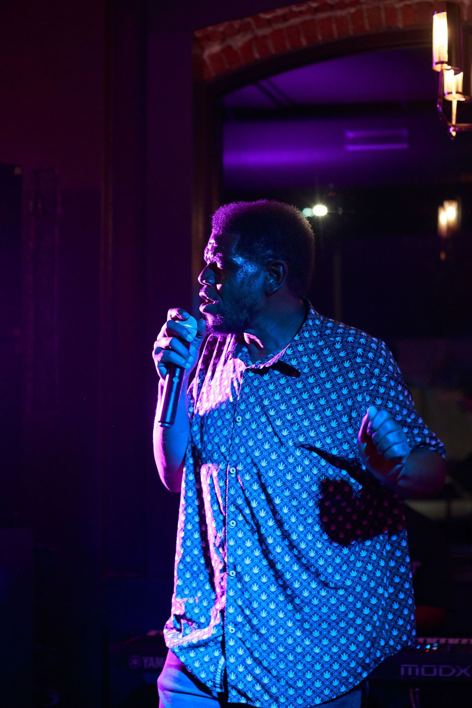 A man in a blue patterned shirt performs on stage holding a microphone under purple stage lighting.