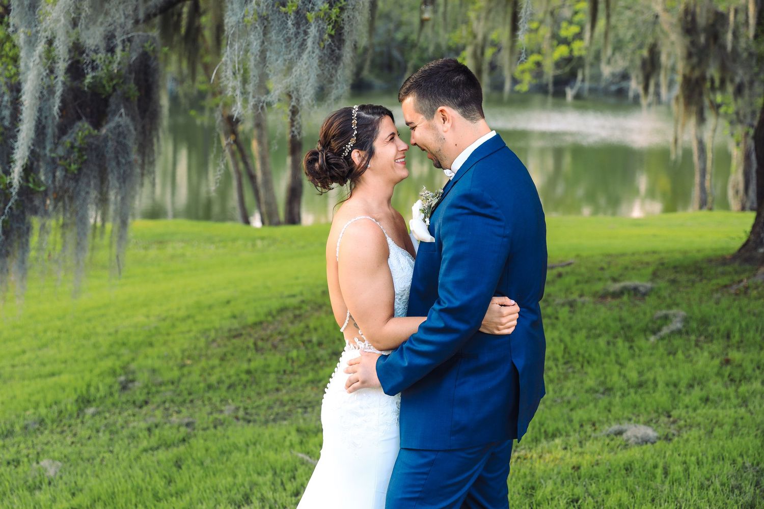 Bride and groom share a romantic embrace outdoors near a pond with Spanish moss-draped trees at their wedding.