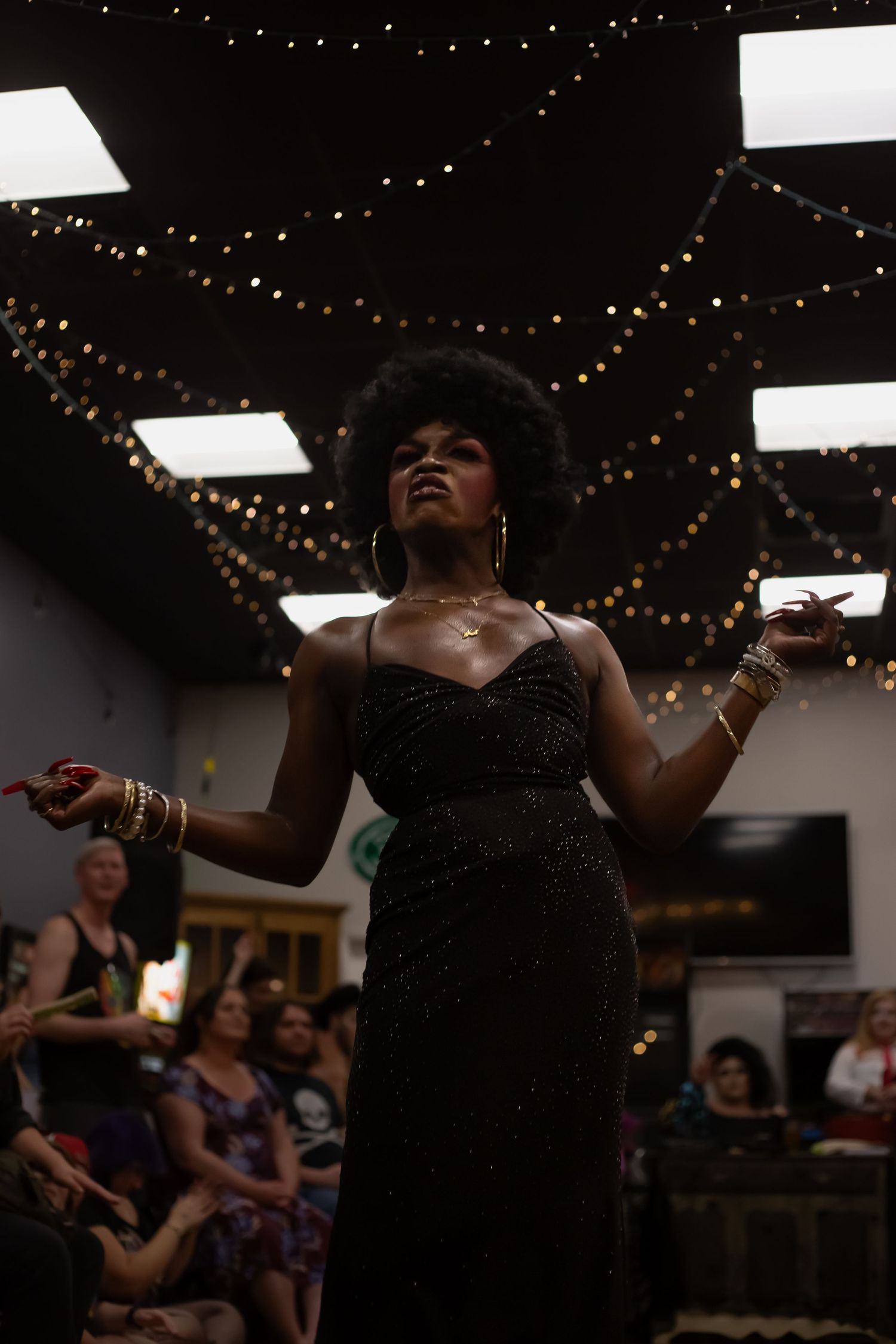 Performer in black gown with afro hairstyle and gold jewelry on stage under string lights at indoor event.