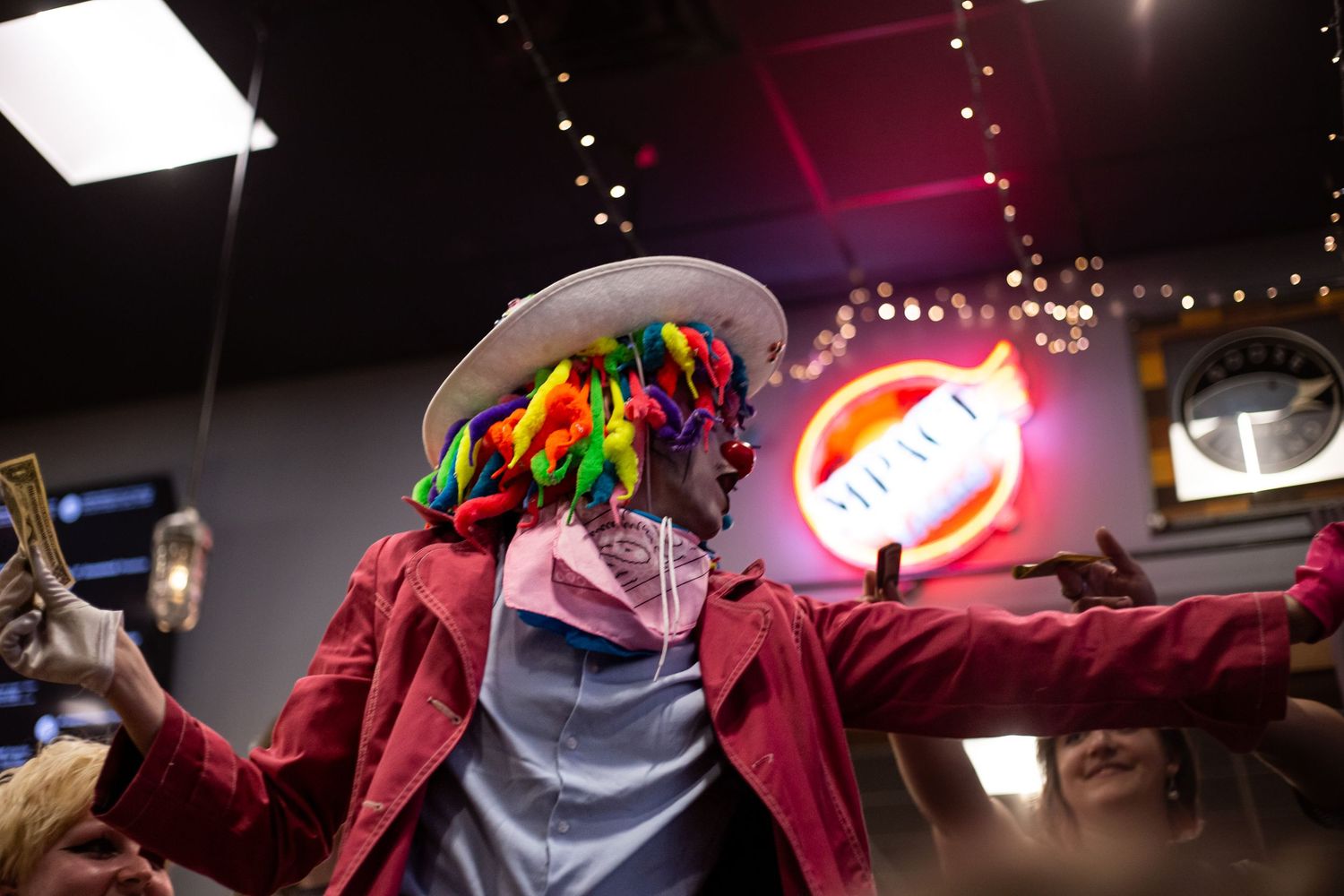 A performer in a colorful clown wig and red jacket dances energetically at a vibrant indoor venue with neon signs.