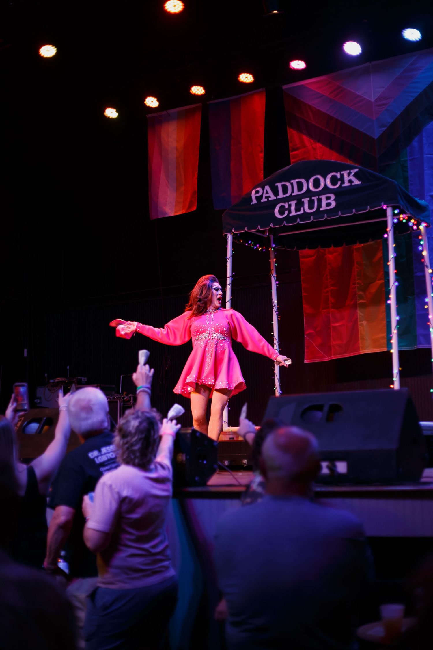 Drag performer in pink mini dress on stage at Paddock Club venue, entertaining crowd under colorful rainbow backdrop.