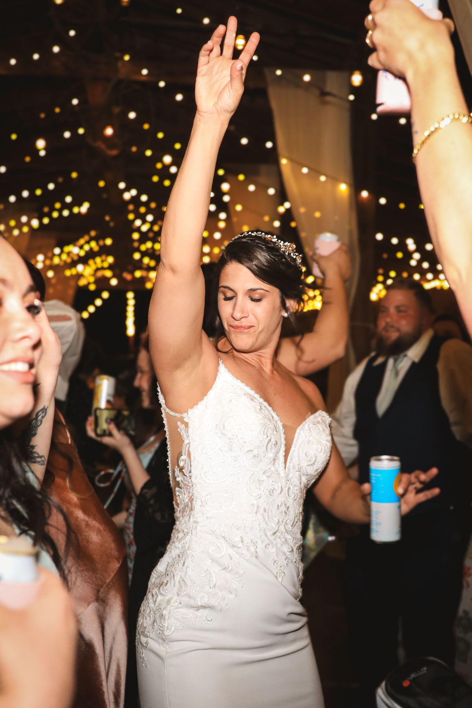 Bride in white lace gown dancing with arm raised at wedding reception with bokeh string lights.