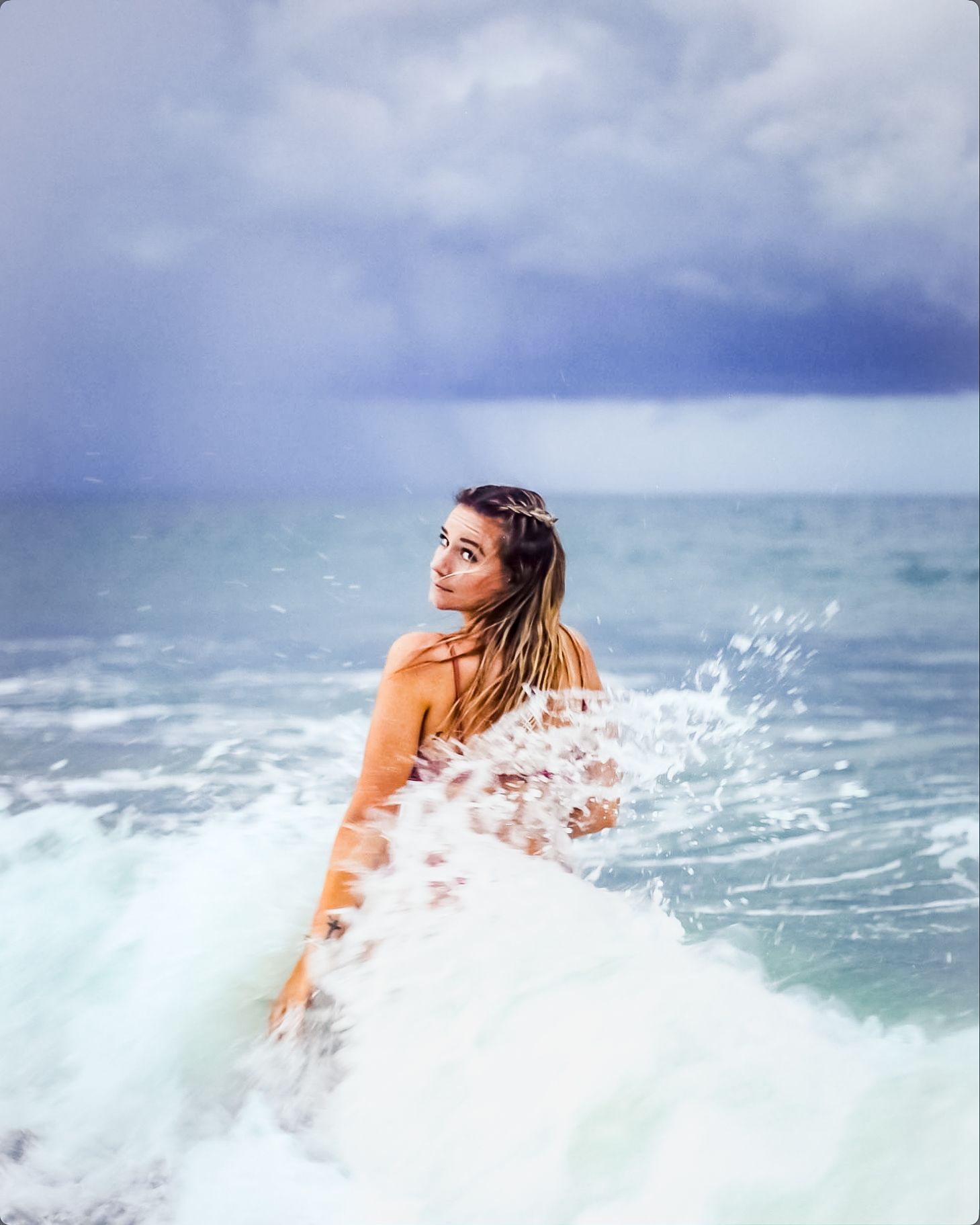 Woman with braids sitting on a surfboard in ocean waves under a dramatic cloudy sky.