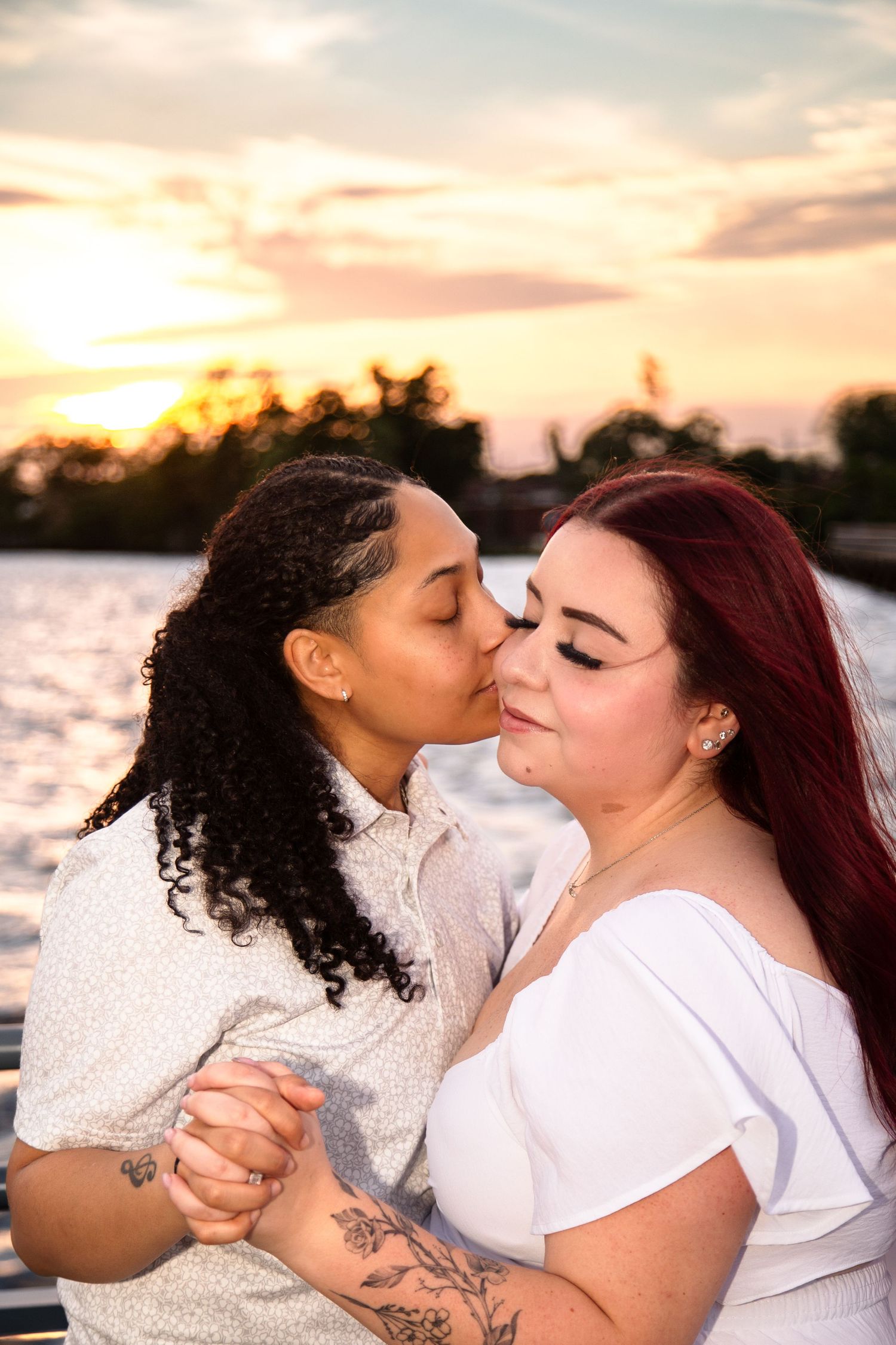 Two women share a romantic kiss at sunset by the water, holding hands, wearing white outfits with tattoos visible.