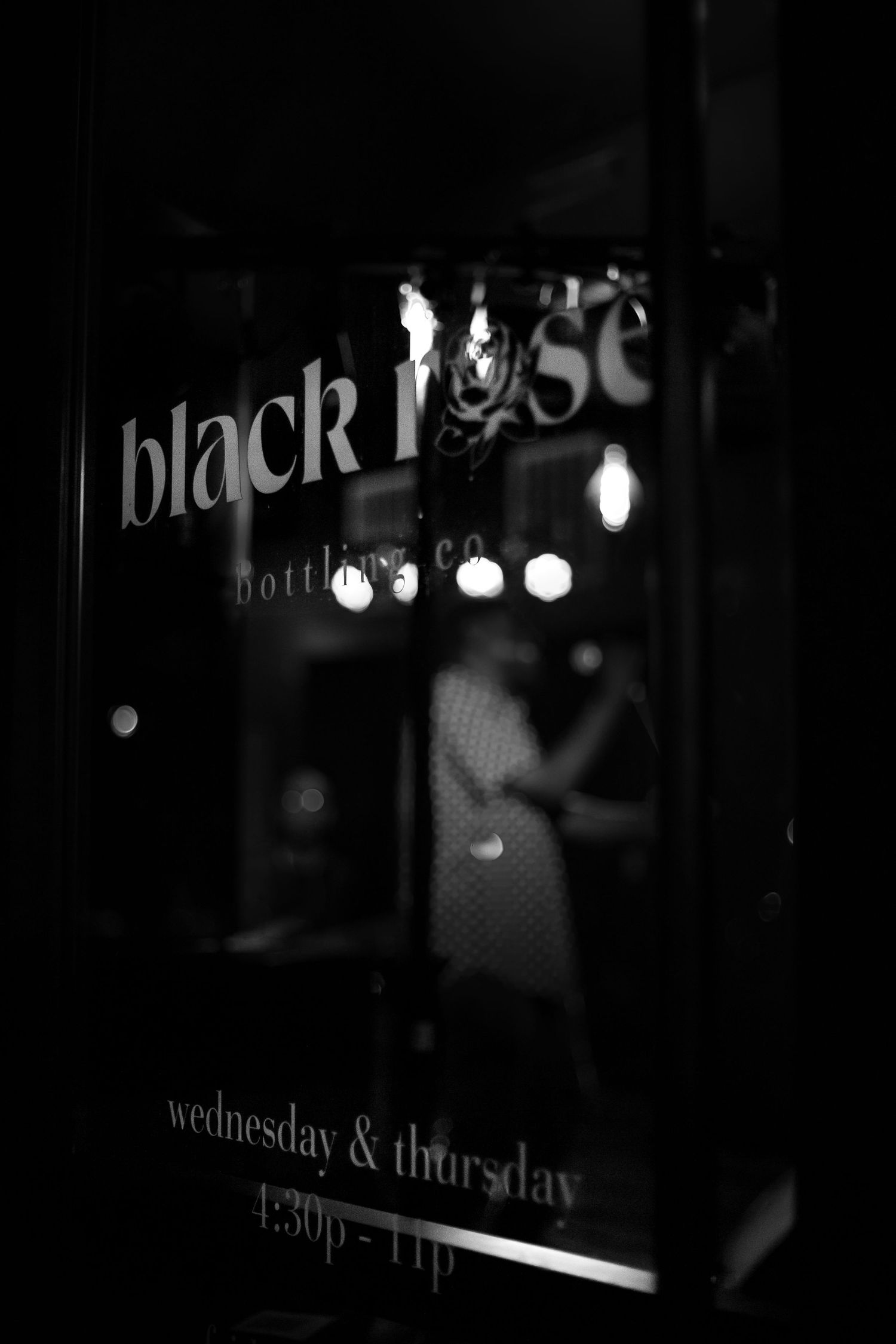 Black and white photo of Black Rose Bottling Co. storefront window showing hours Wednesday & Thursday 4:30p-11p.