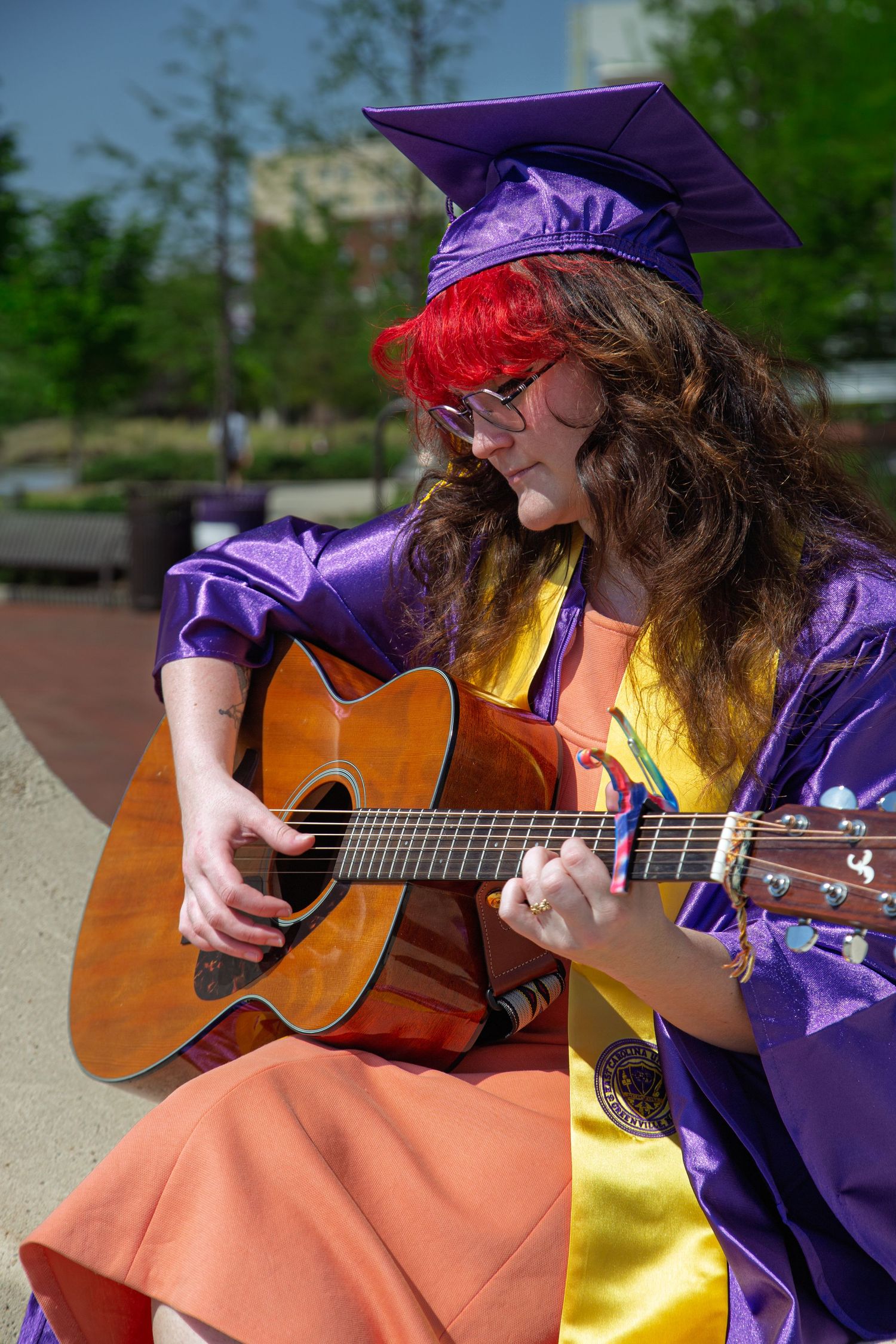 Graduate in purple cap and gown plays acoustic guitar outdoors, wearing red headband and colorful attire.