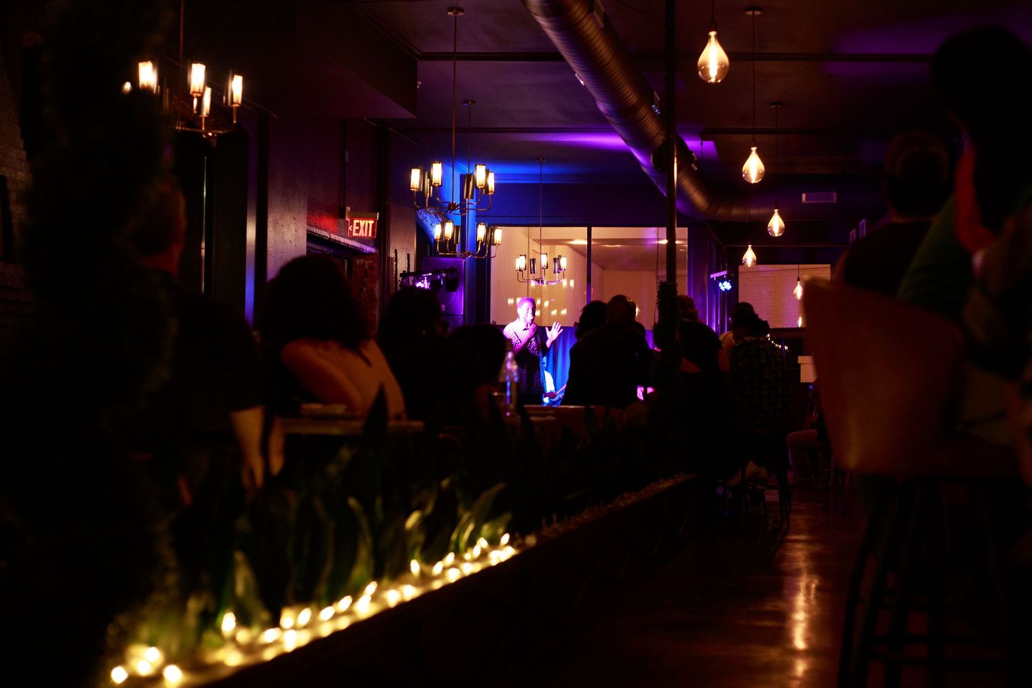 Dimly lit bar lounge with blue and purple ambient lighting, silhouetted guests, and glowing candles along a wooden bar.