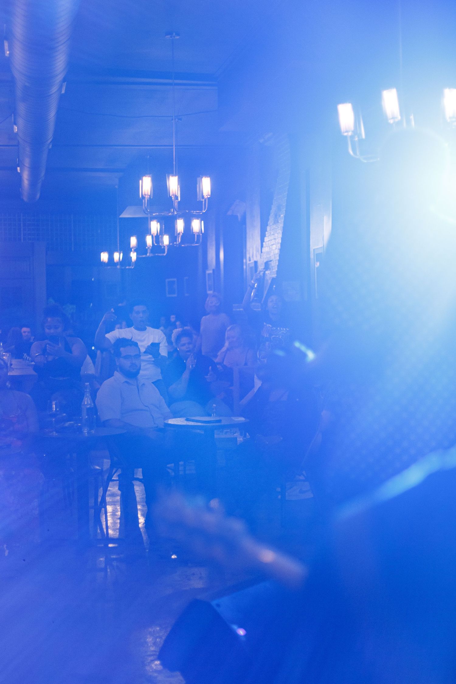 Blue-lit nightclub venue with audience seated at tables watching a live performance on stage.