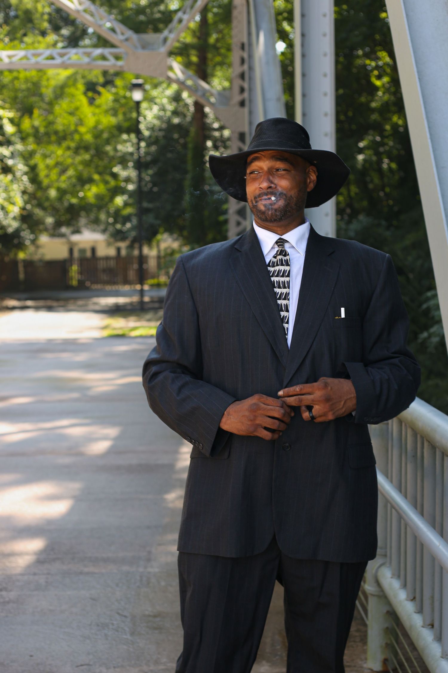 Confident Black man in dark suit, patterned tie, and wide-brim hat standing on a bridge surrounded by trees.