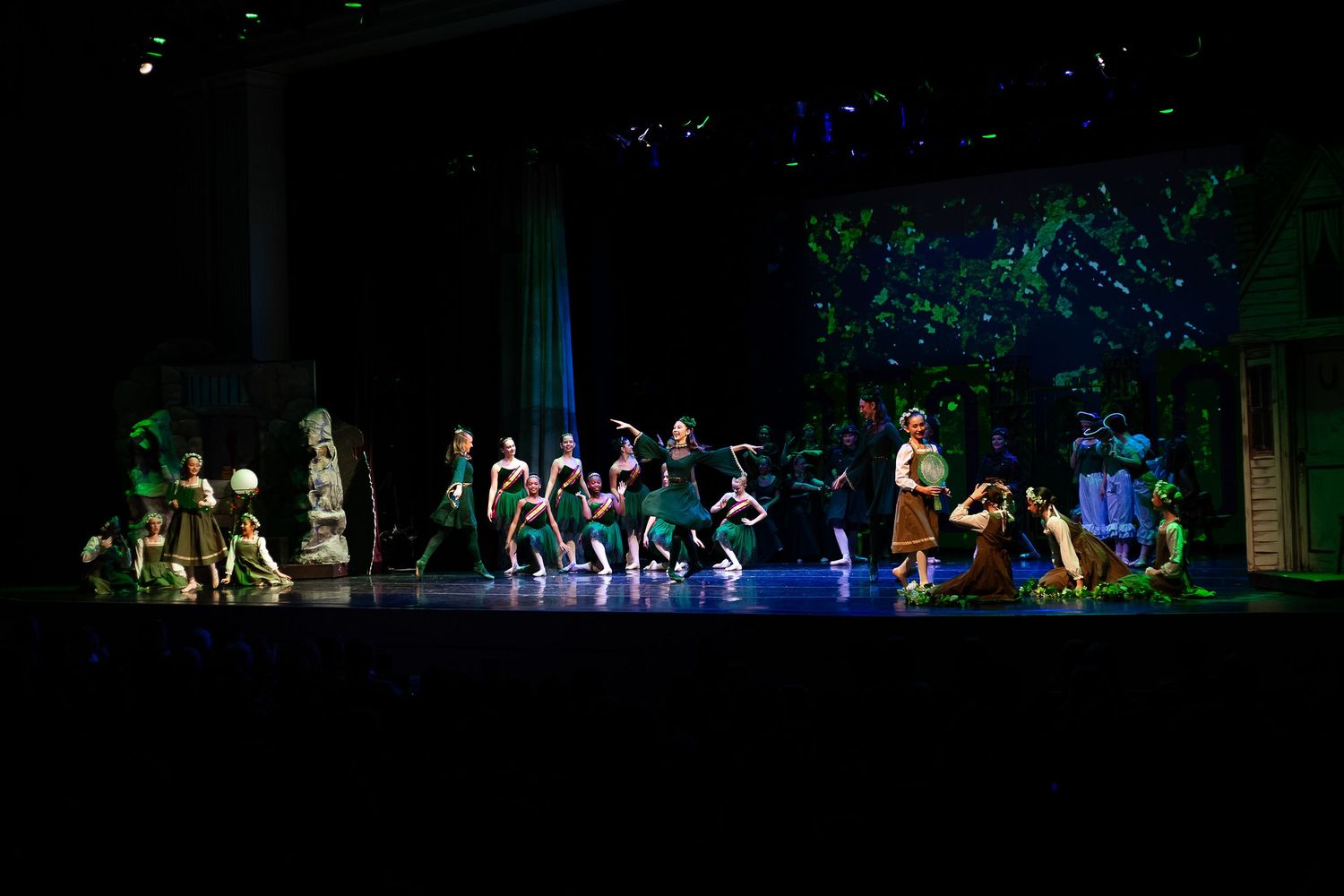 Theater performers in green costumes on a dramatic stage set with trees and blue lighting during a live production.