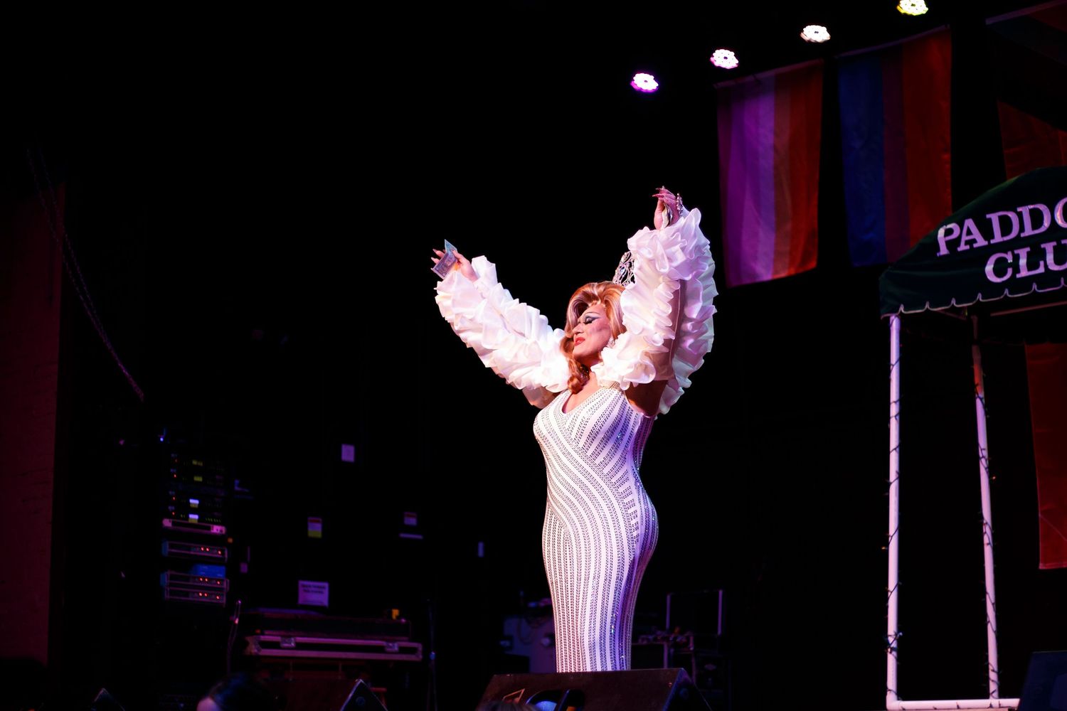 A performer in a white sequin dress with feathered wings poses on stage at the Paddock Club.