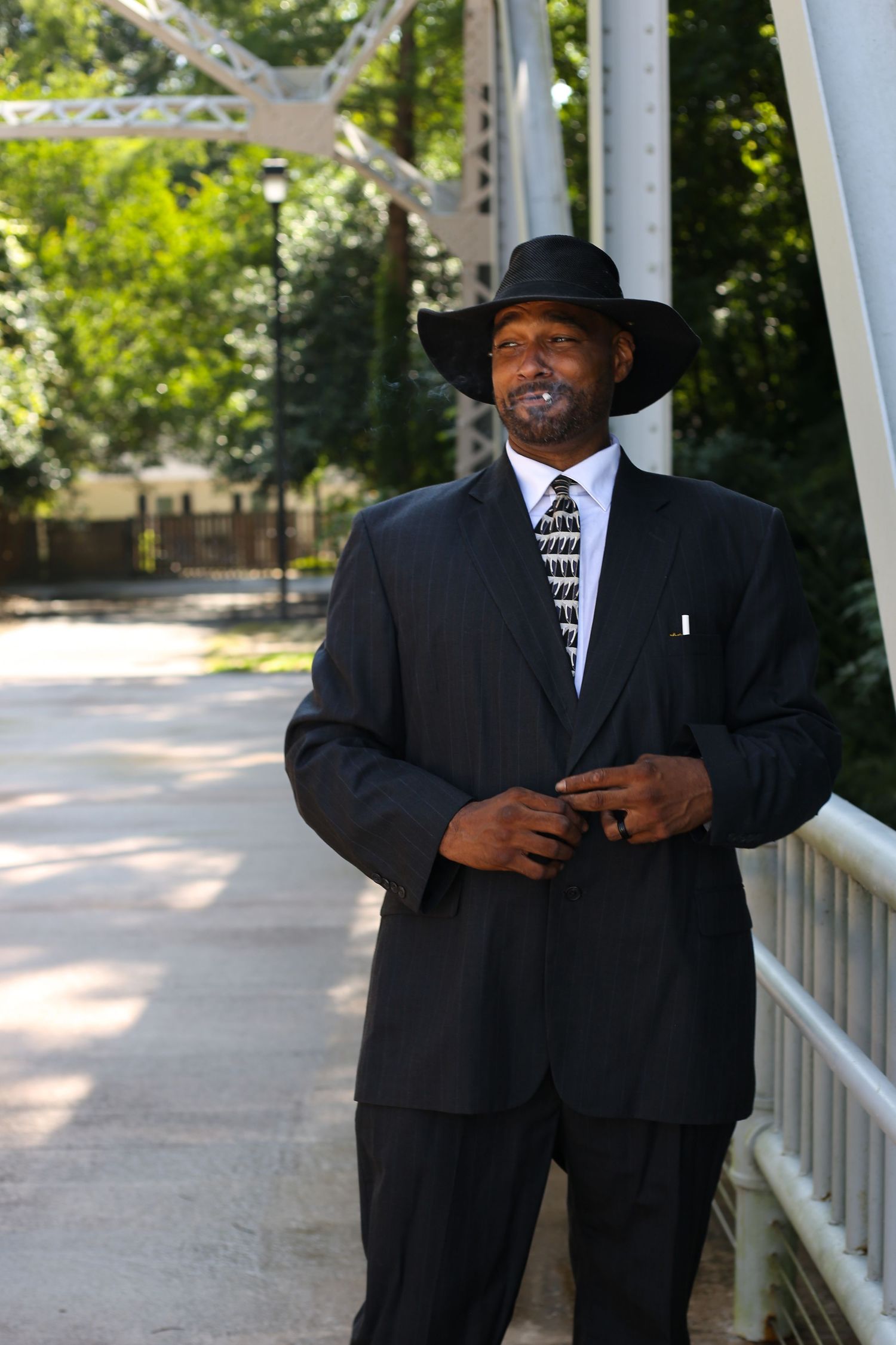 A well-dressed Black man in a dark suit, patterned tie, and wide-brim hat stands confidently on a bridge outdoors.