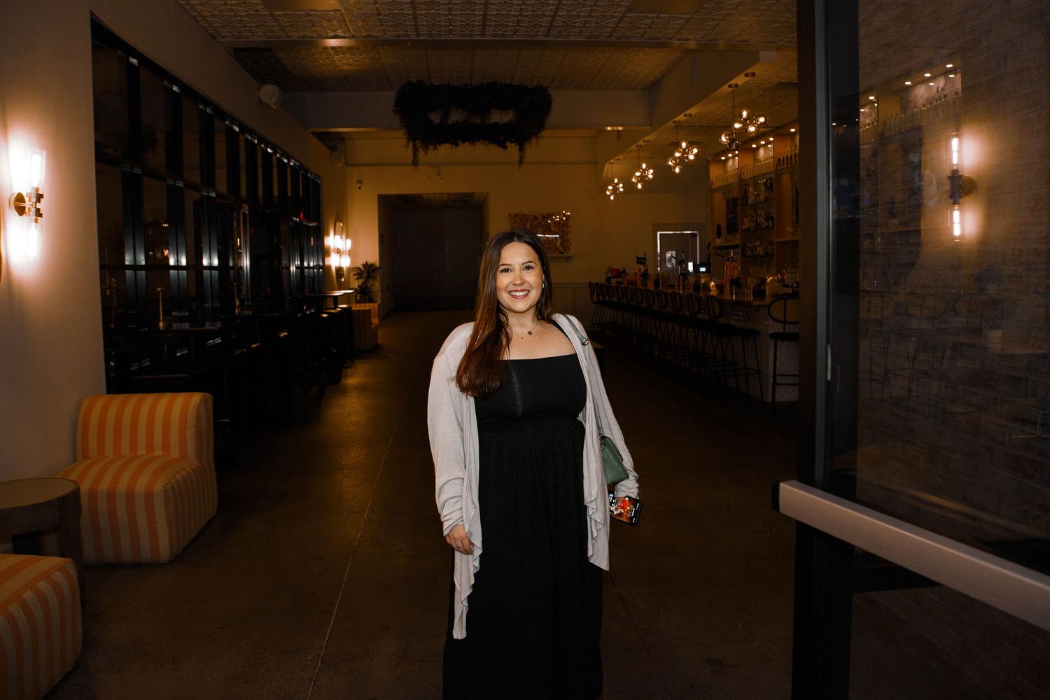 Woman in black dress and white cardigan smiling in a dimly lit upscale bar or event venue.