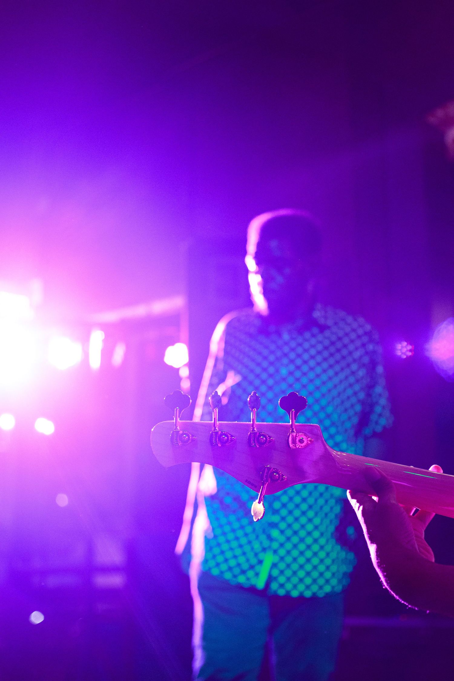 Musician playing bass guitar on stage with vibrant purple and pink concert lights glowing in the background.