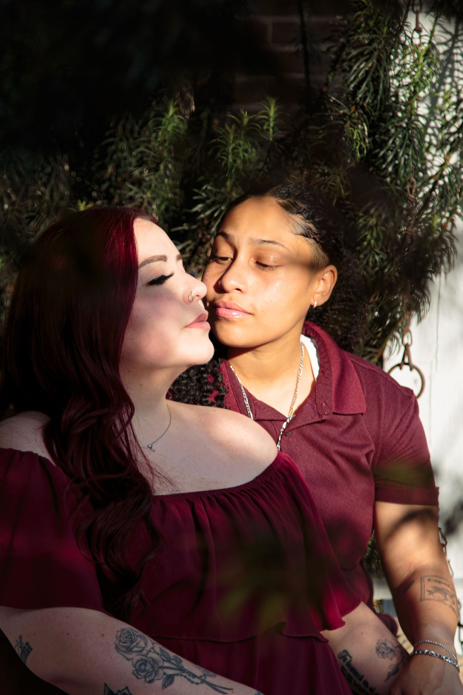 Two women in matching burgundy outfits sharing a kiss outdoors.
