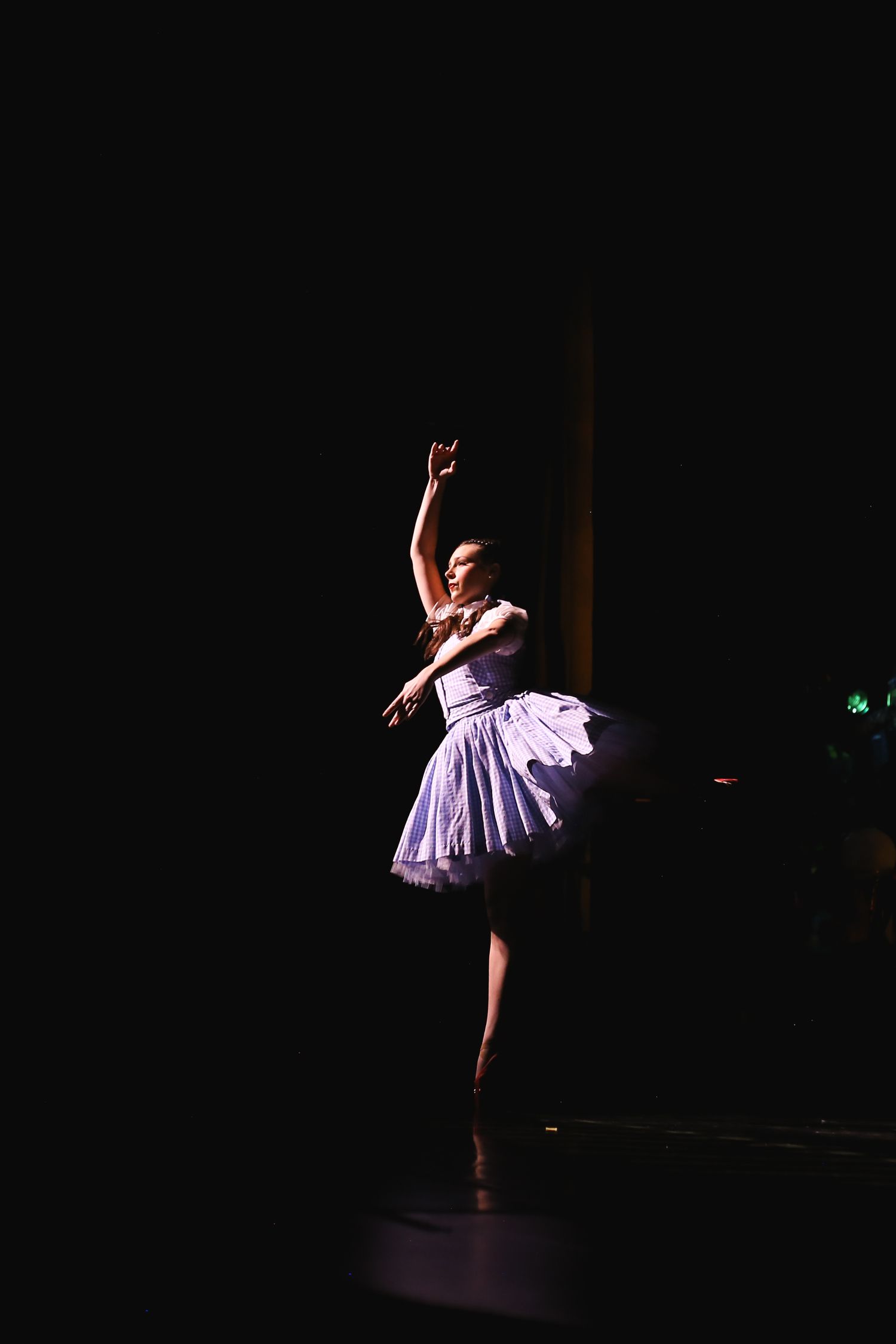 A ballerina in a purple tutu performs gracefully on a dimly lit stage with one arm raised overhead.