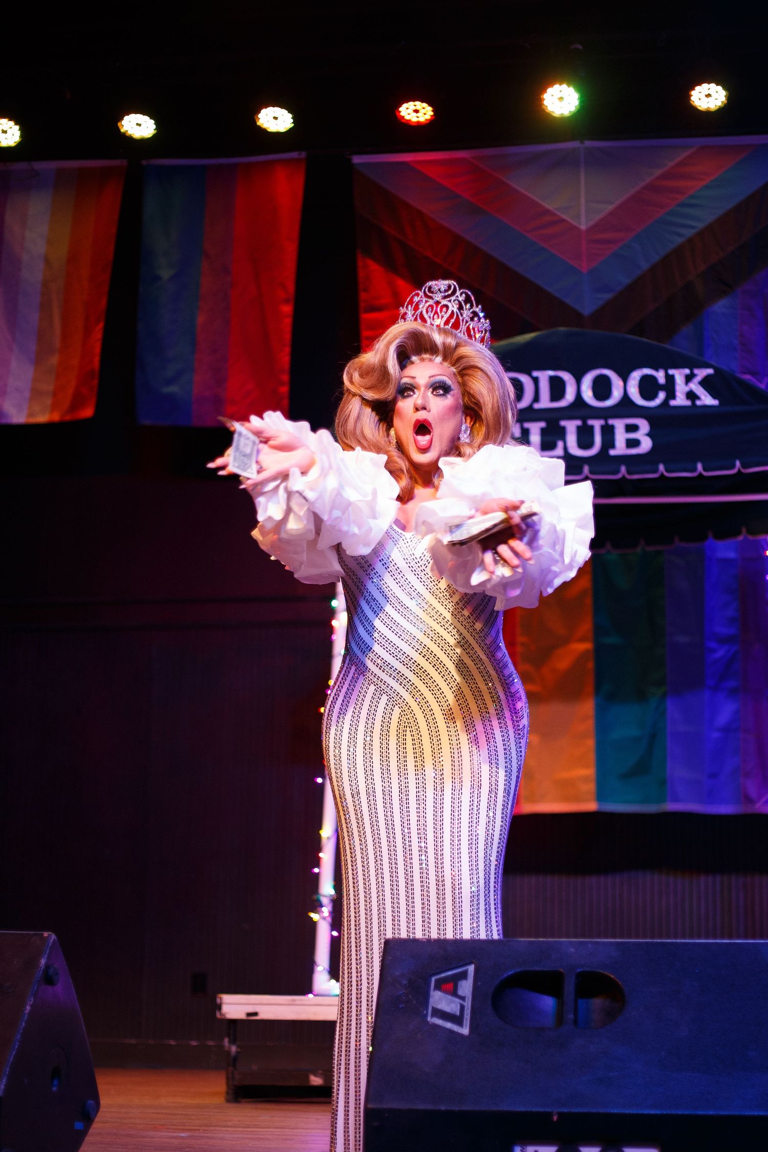 Drag queen performer in sequined gown and tiara on stage with rainbow pride flags at the Paddock Club.