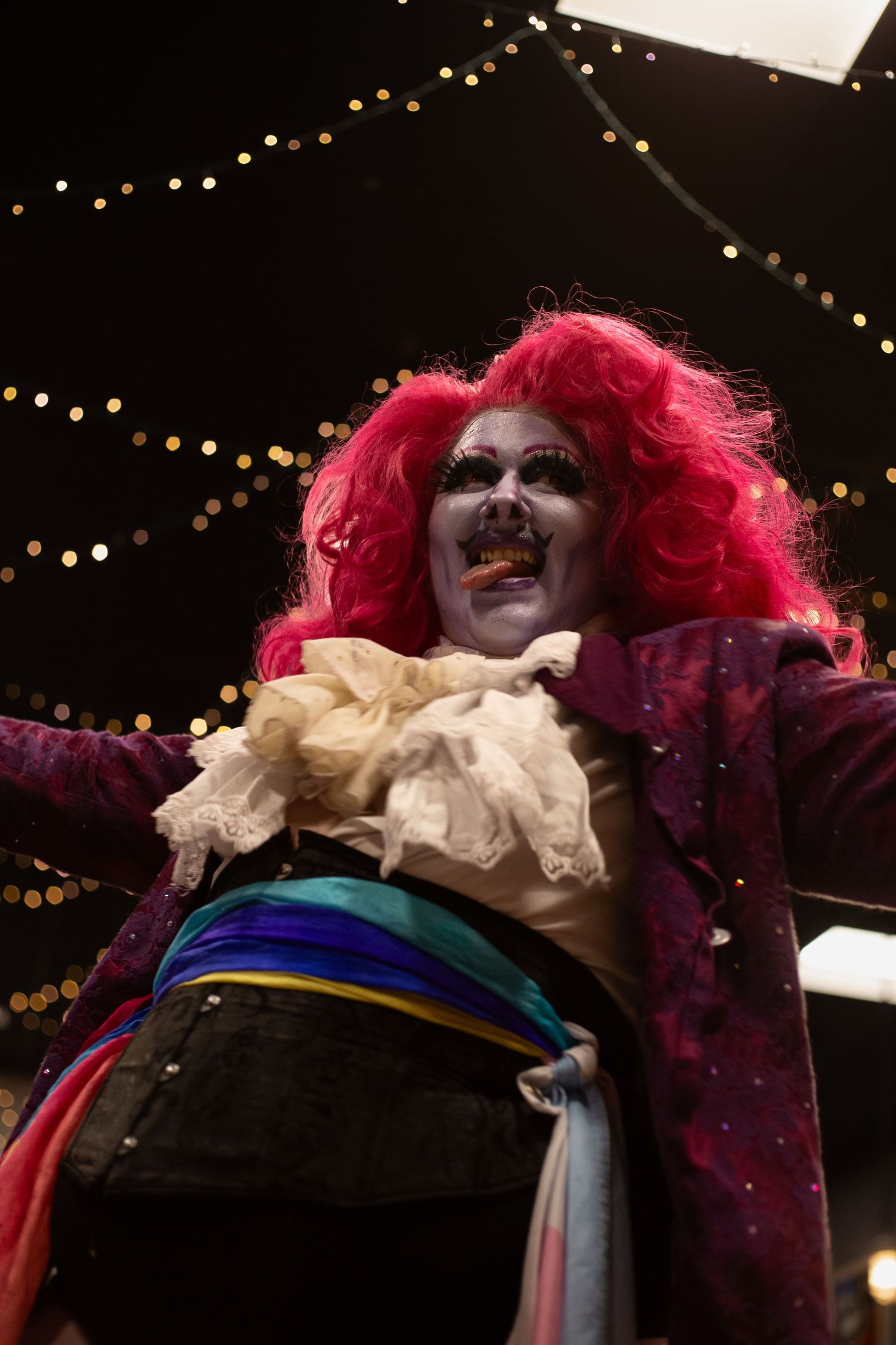 Dramatic clown performer with bright pink wig, white face paint, rainbow sash, and purple costume under string lights.