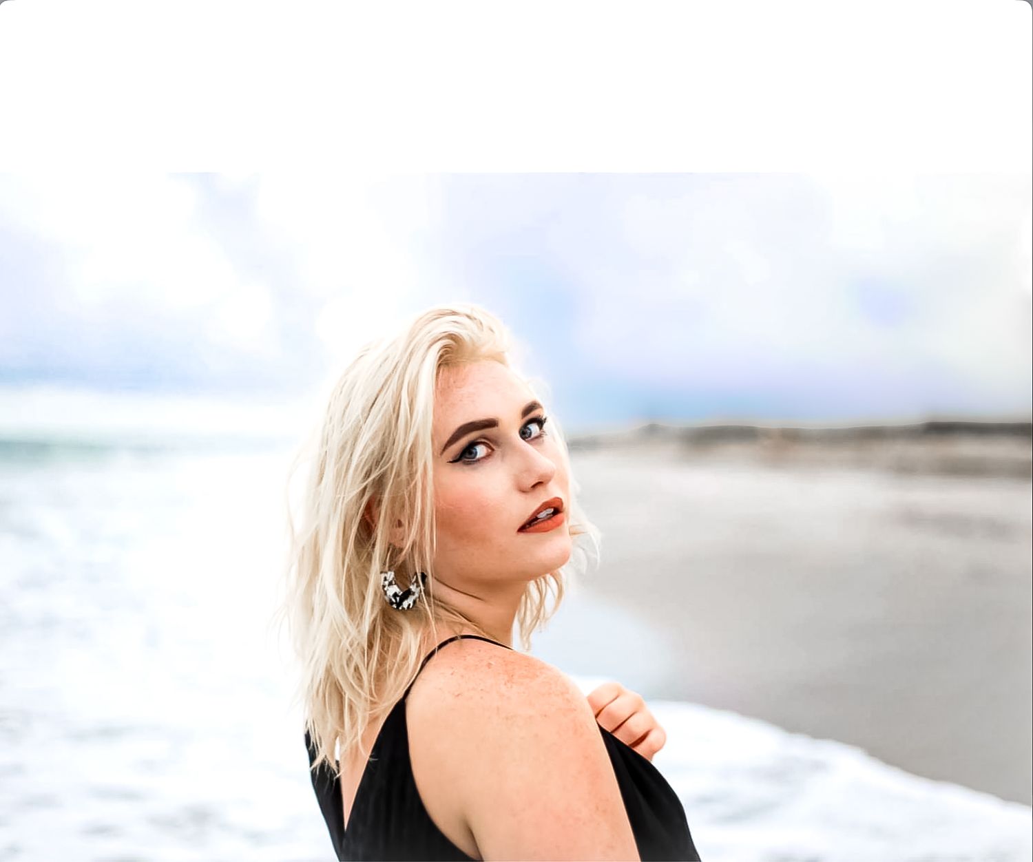 Blonde woman in black dress posing on a beach with ocean waves and cloudy sky in the background.